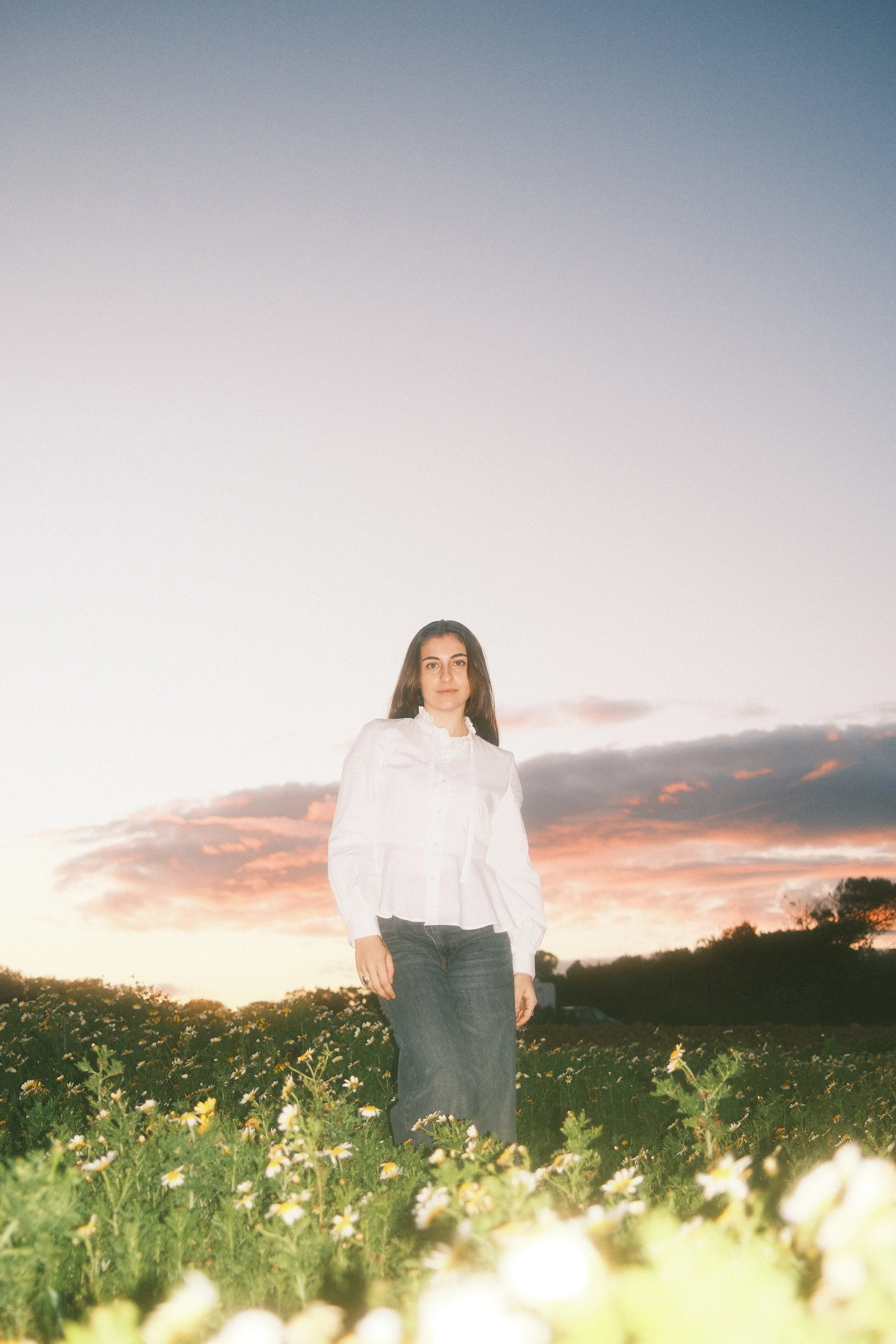 A young woman standing in a field with daisies during sunset, wearing a white blouse and dark skirt.