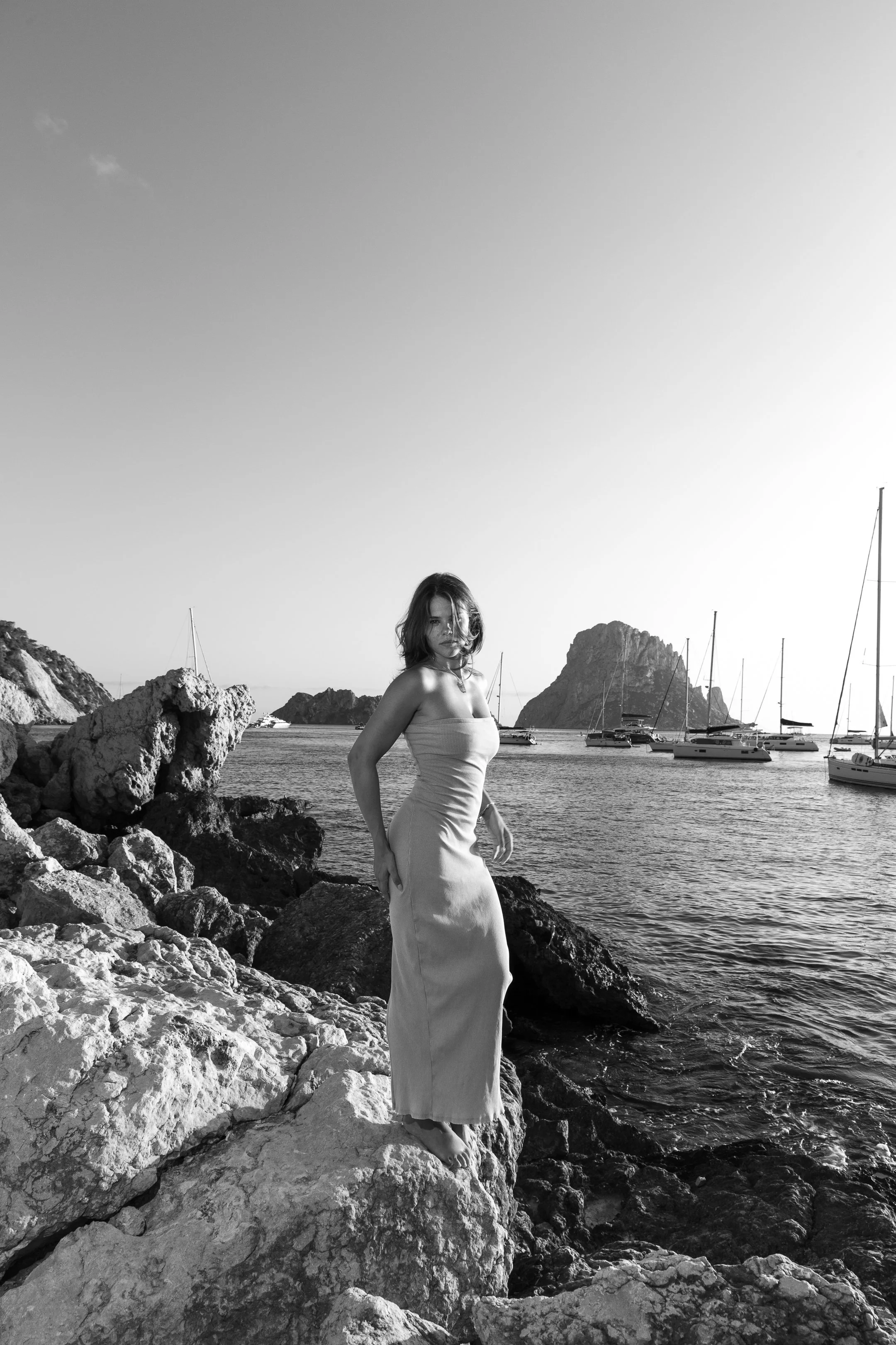 A woman in a strapless dress standing on rocks at the edge of water with sailboats and a mountain in the background, black and white photo.