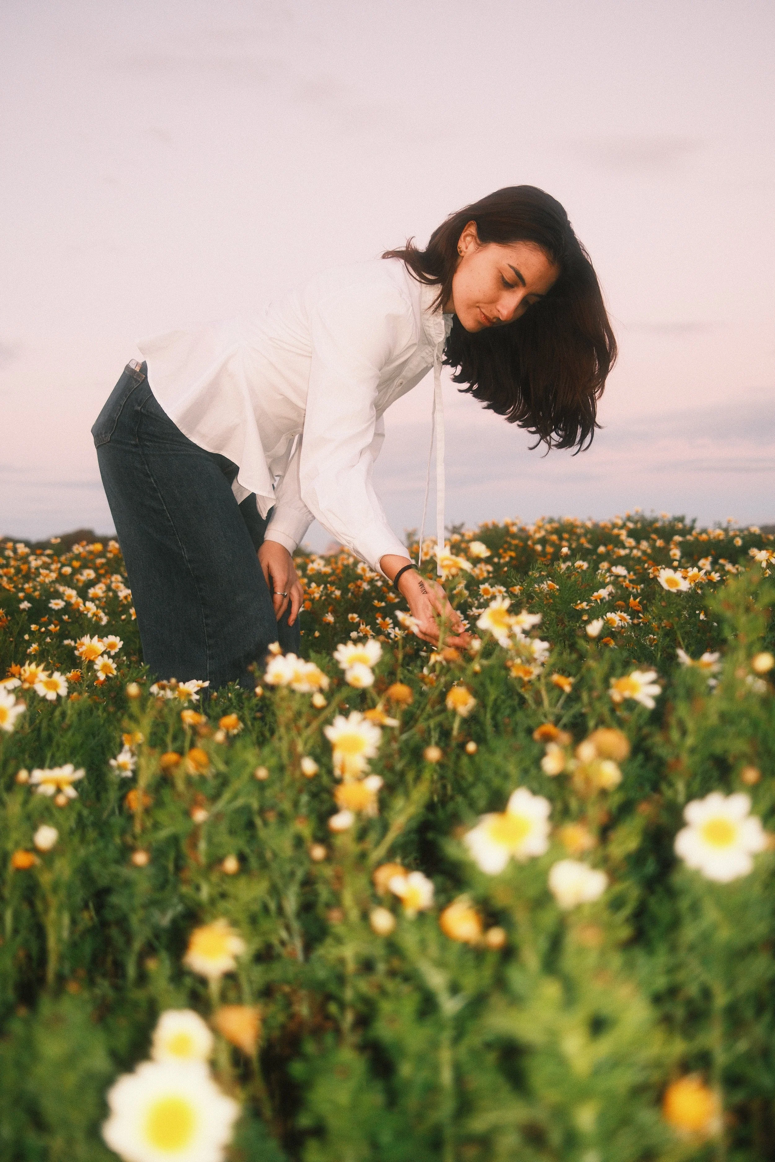 A woman with shoulder-length dark hair, wearing a white shirt and dark jeans, is bending down and touching yellow and white daisies in a field during sunset or sunrise.