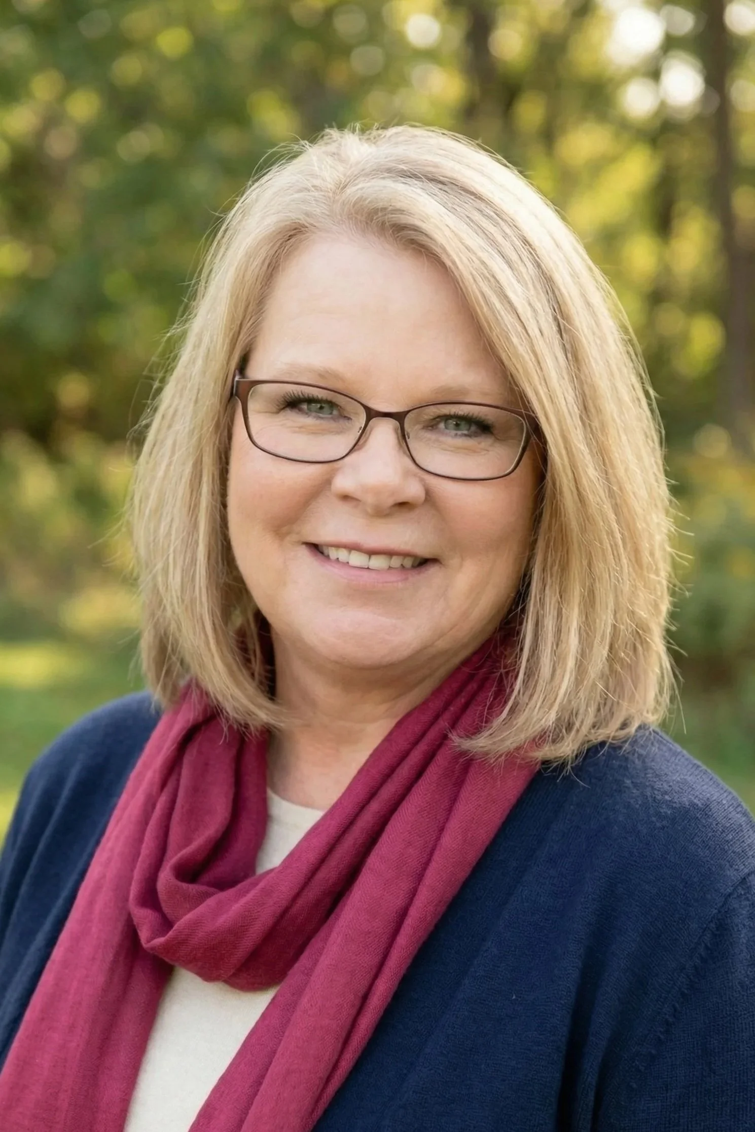 A intuitive mentor with blonde hair, wearing glasses, a blue blazer, and a red scarf, smiling outdoors with a green, leafy background.