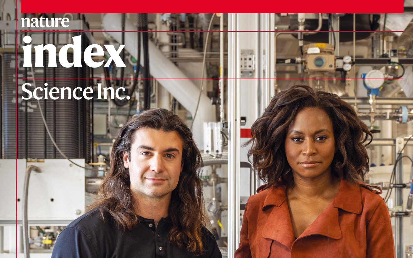 Two scientists, a man and a woman, standing in a laboratory with complex equipment and pipes in the background.