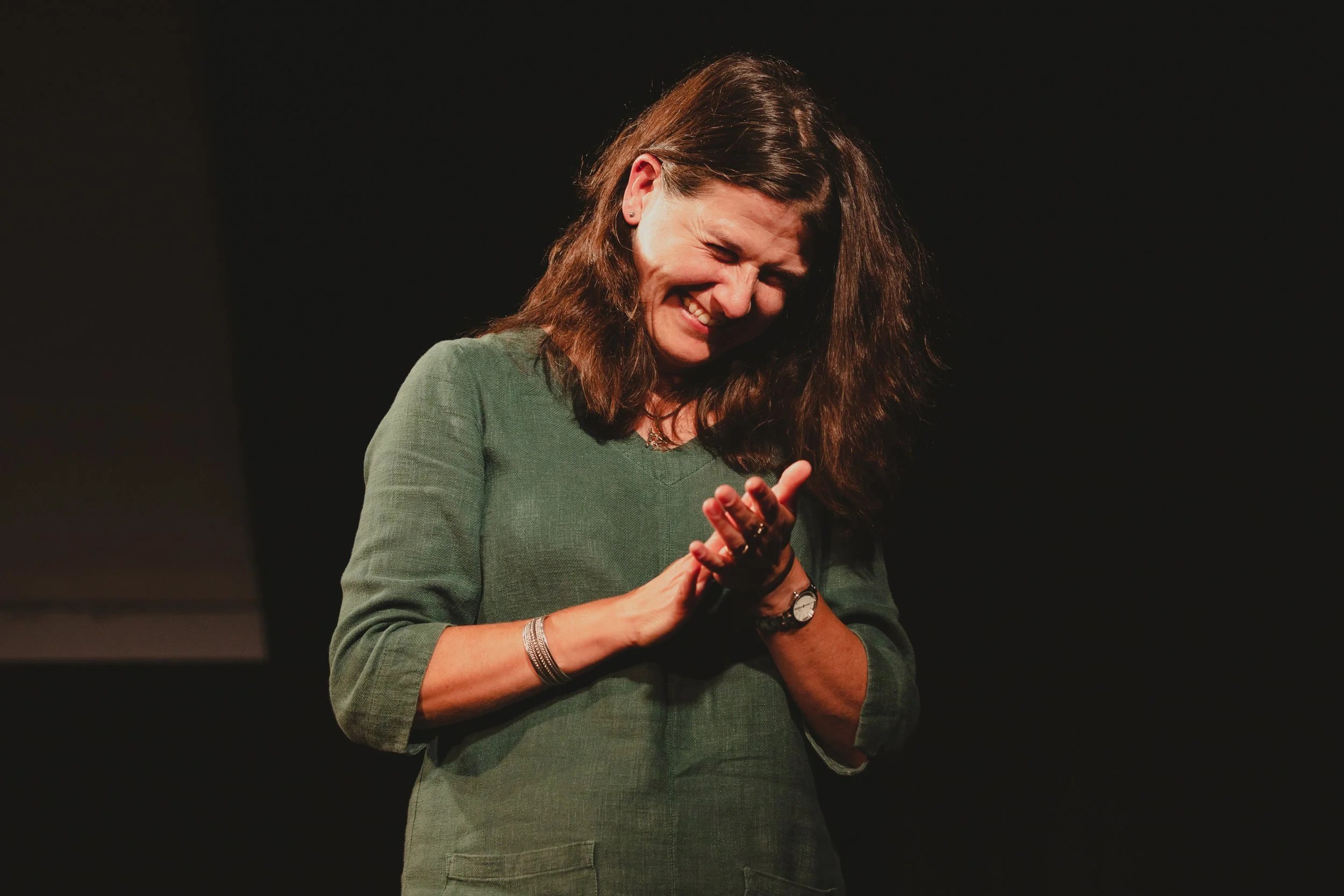 Lisa Schneidau, wearing a green top, smiling and laughing while looking down, on a dark stage background during a performance.