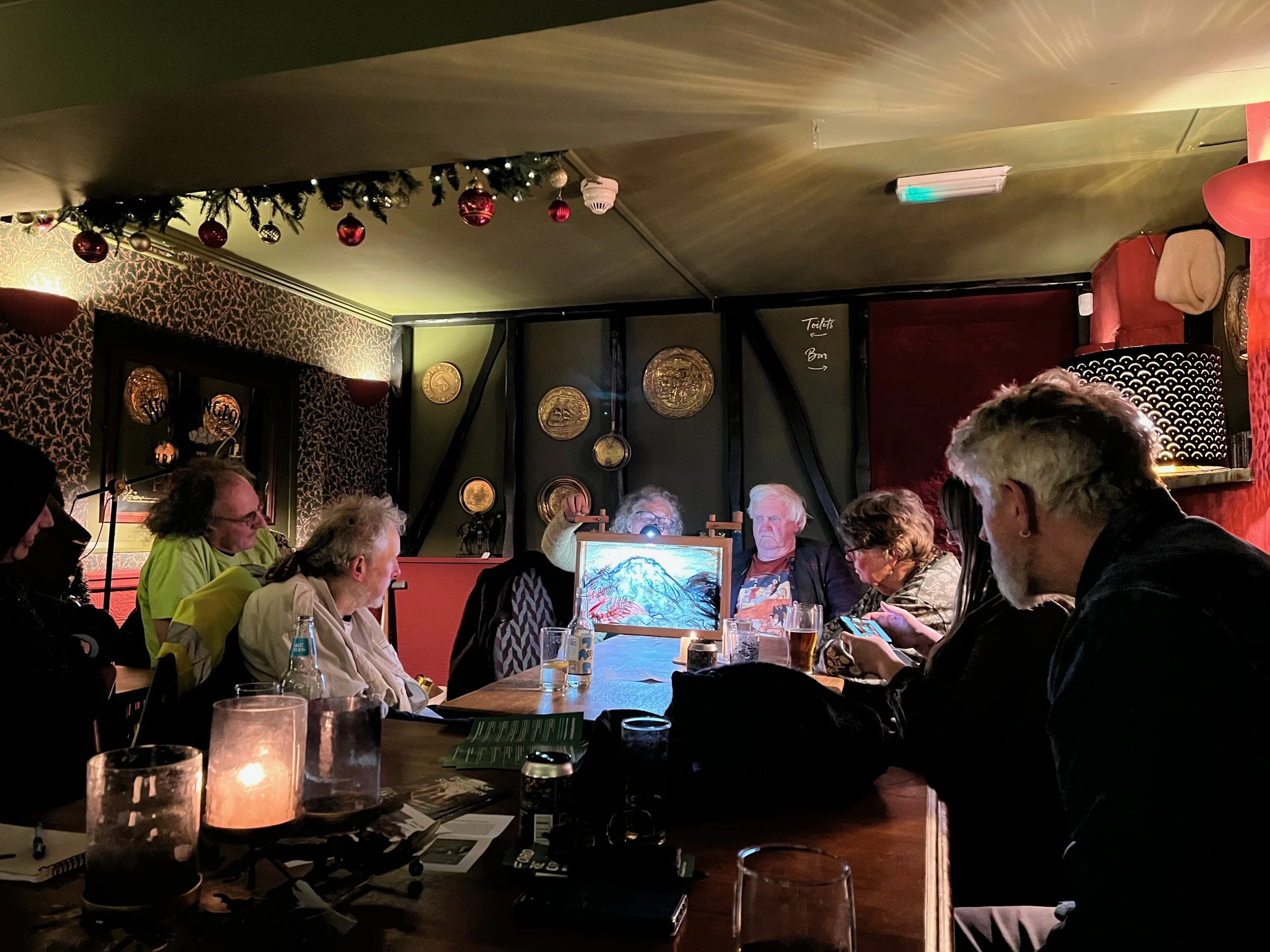 A group of people seated at tables in a cozy pub with festive holiday decorations, including red and gold ornaments hanging from the ceiling and framed gold wall art, during a storytelling session.