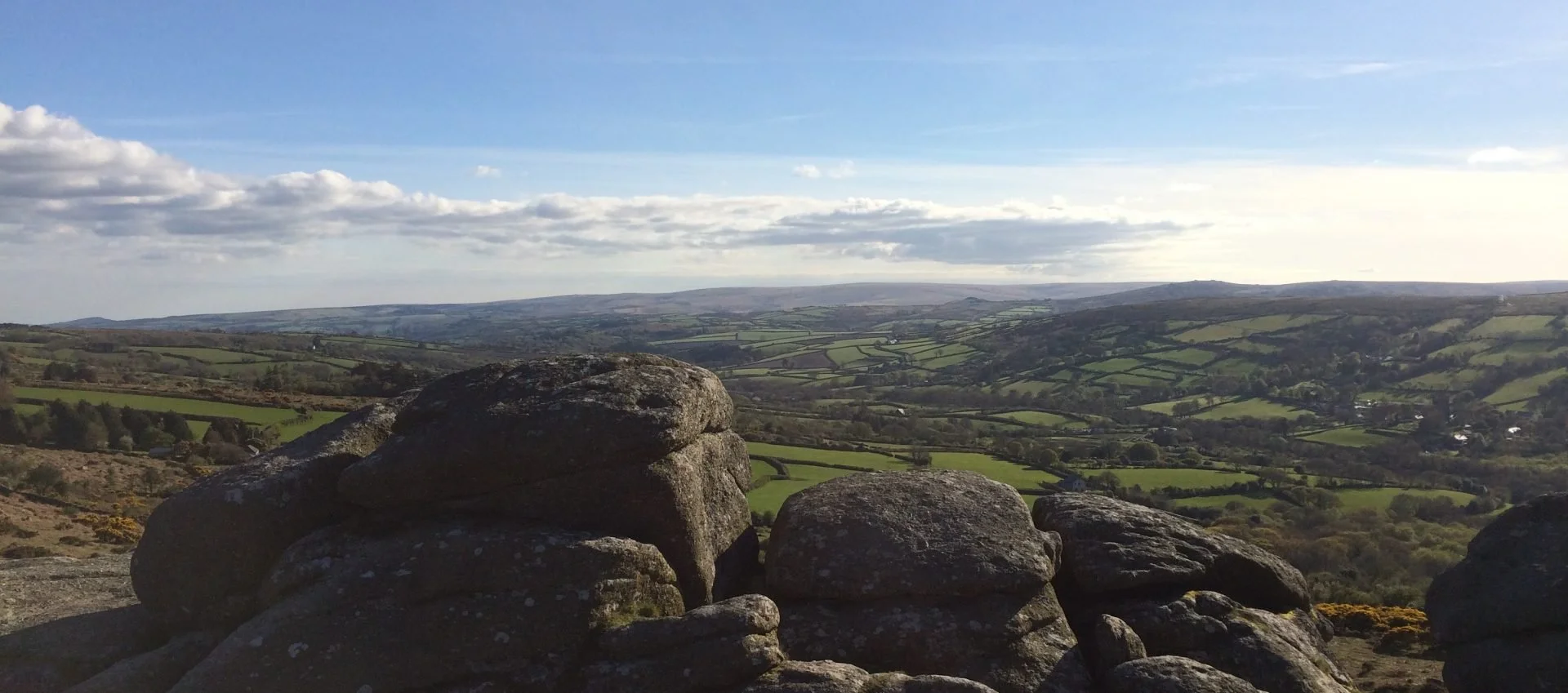 A scenic view of the rolling green hills and fields of Dartmoor under a partly cloudy sky.