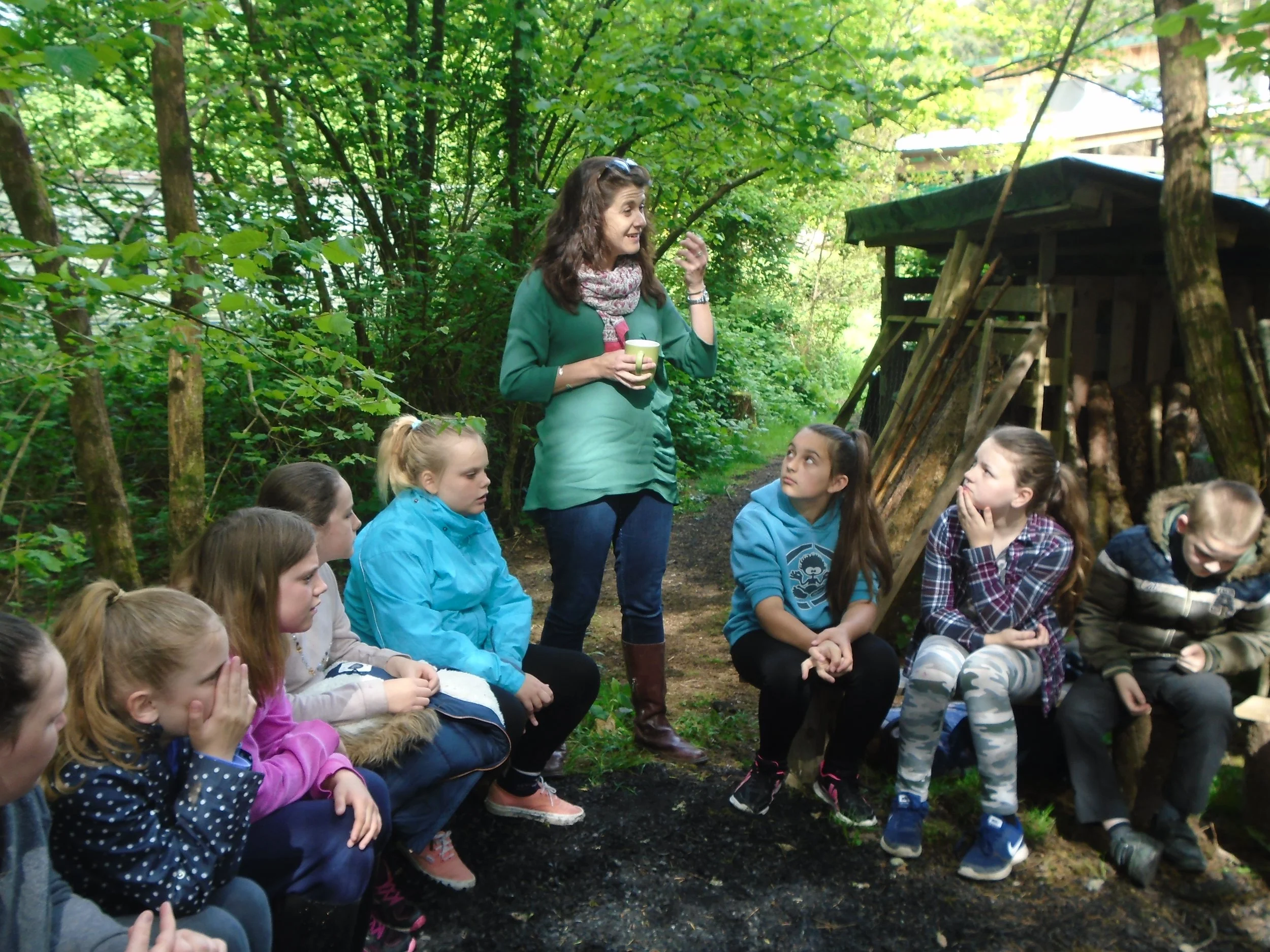 Lisa Schneidau holding a mug and speaking to a group of children sitting in a semicircle outdoors in a wooded area.