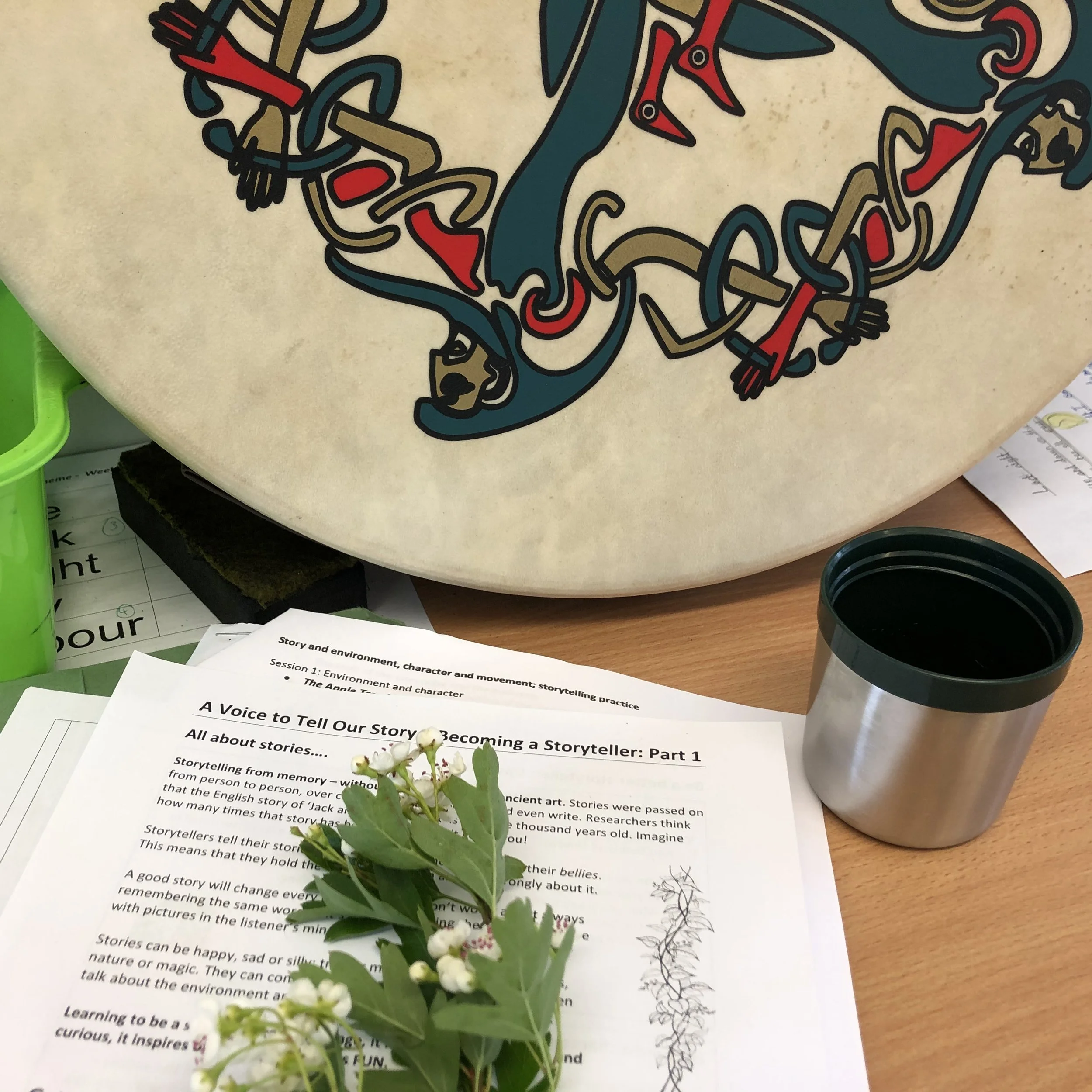 A close-up photo of a small flowering plant with green leaves and white buds, placed on a desk in front of printed papers. The papers contain text about storytelling and becoming a storyteller.
