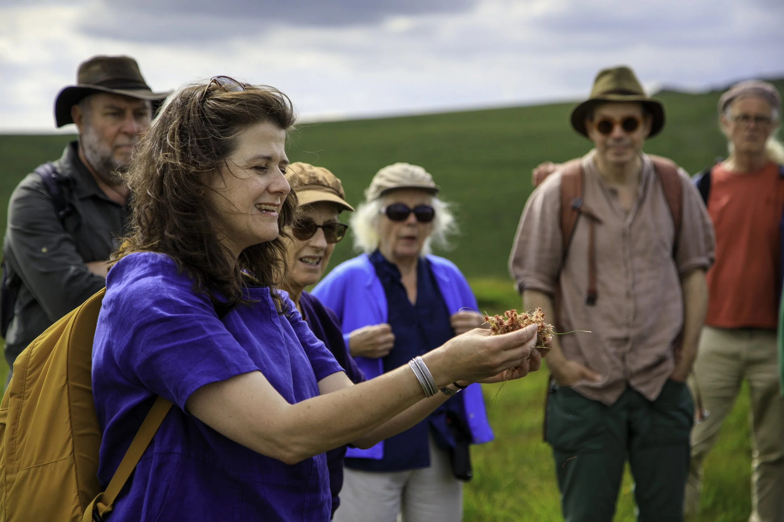 Lisa Schneidau is with a group of people outdoors, standing on a grassy hill with overcast sky.