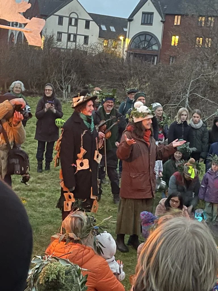 Group of people gathered outdoors in front of residential buildings during dusk. Some are dressed in costumes, including a woman in a witch costume with orange decorations and a hat, and another woman with a leafy wreath on her head. Others are dressed casually, some smiling and watching the scene.