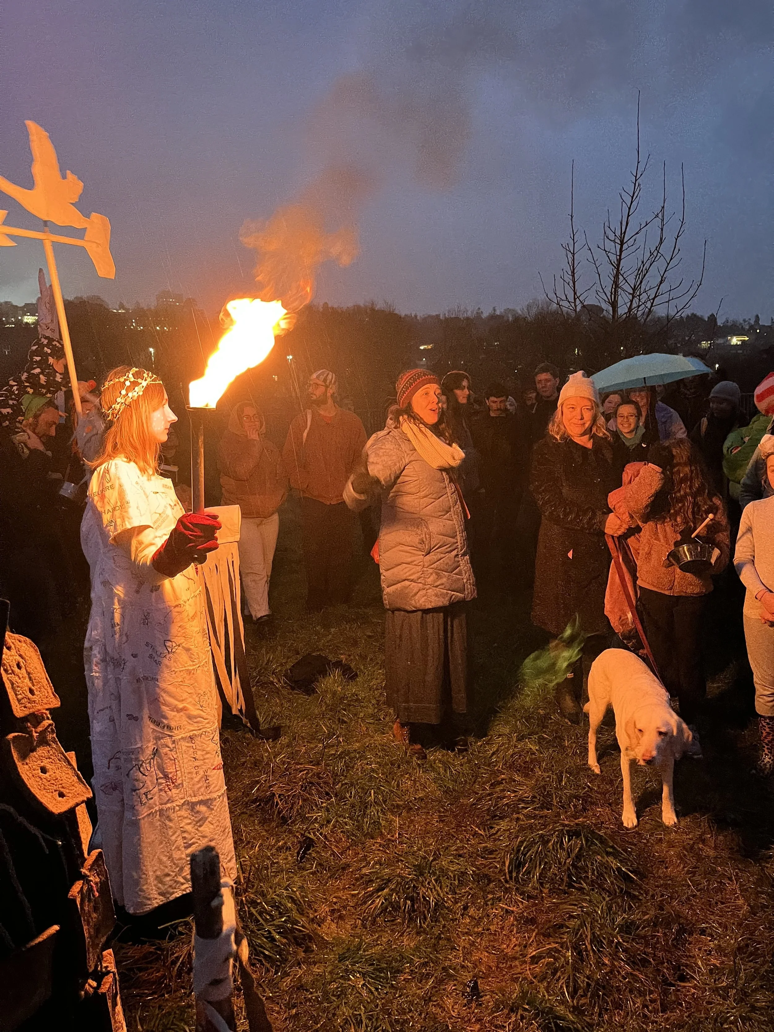 A group of people, some with umbrellas, gather outdoors near a fire with smoke rising into the sky during dusk or evening.
