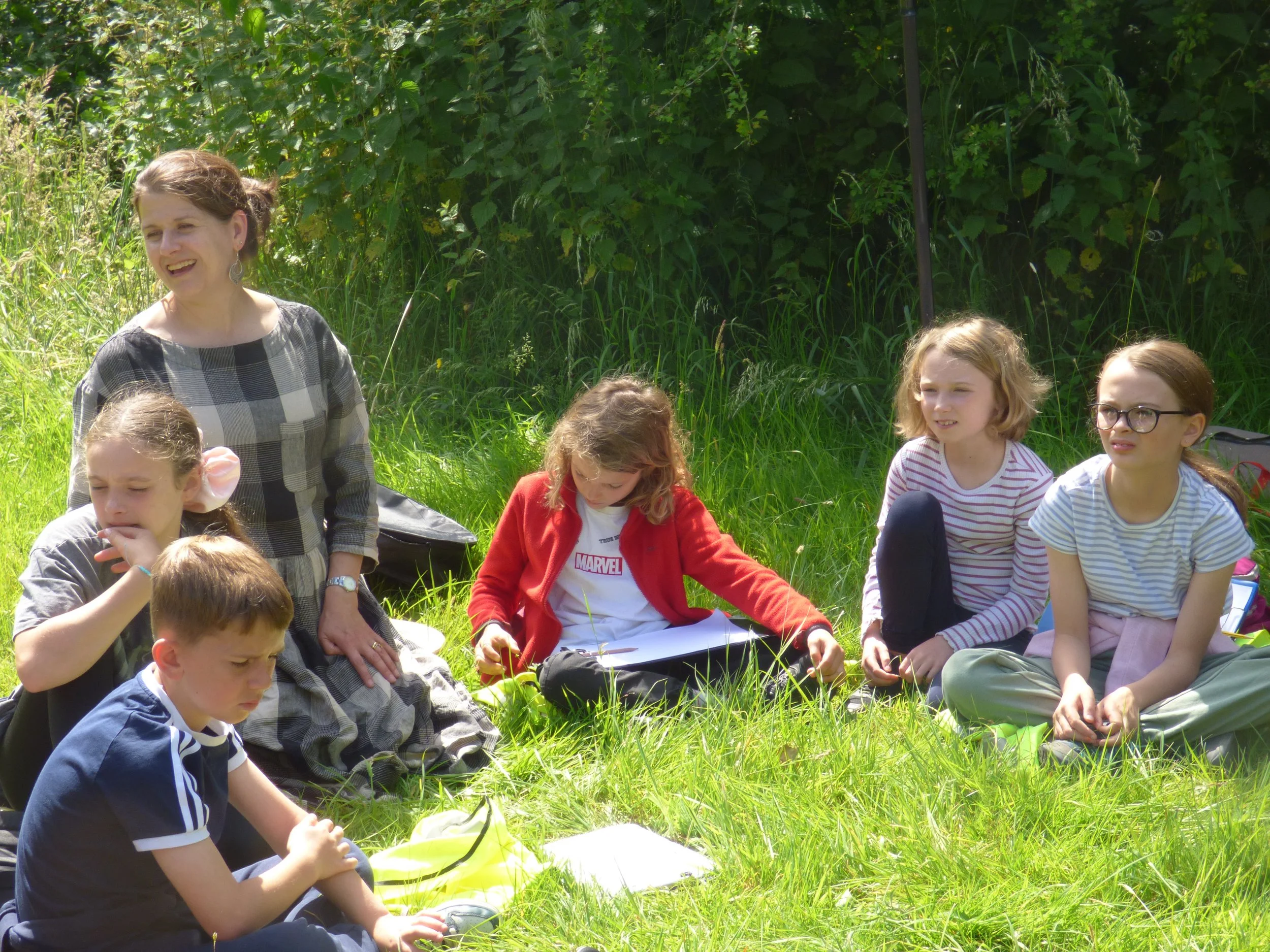 Lisa Schneidau outdoors with a group of children sitting on the grass. The children are engaging in an outdoor activity, with some looking at a notebook or paper and others resting or listening. The background shows dense green foliage and plants.