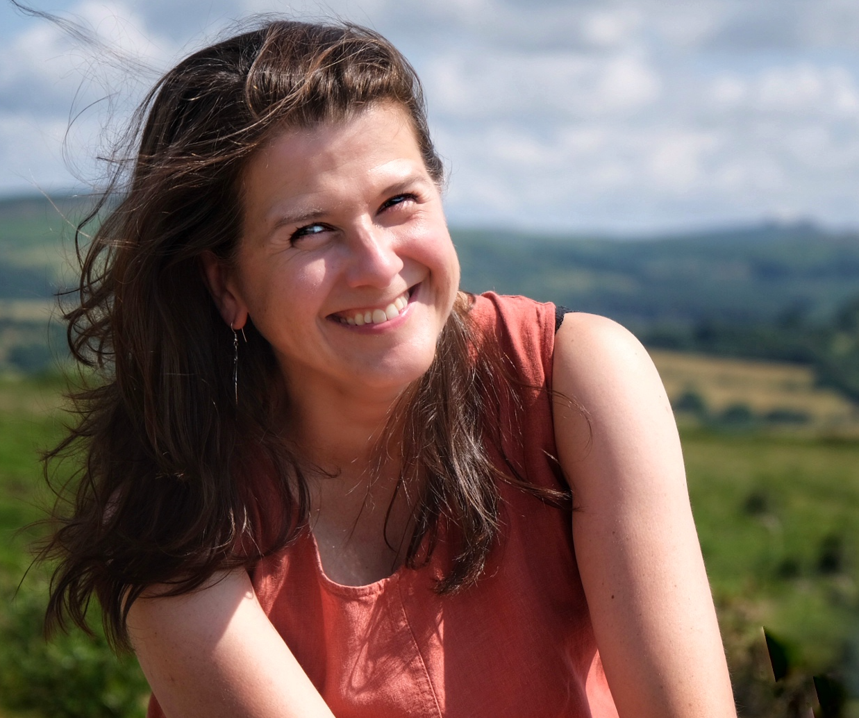 Lisa Schneidau is smiling outdoors with the green hilly landscape of Dartmoor and cloudy sky in the background.