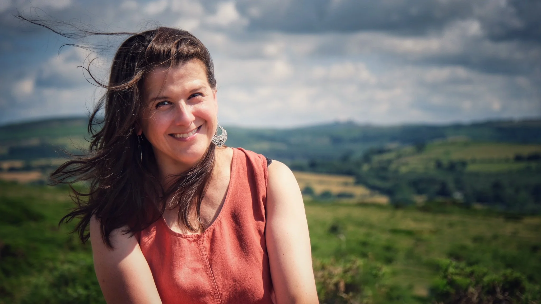 Lisa Schneidau is smiling outdoors with the green hilly landscape of Dartmoor and cloudy sky in the background.