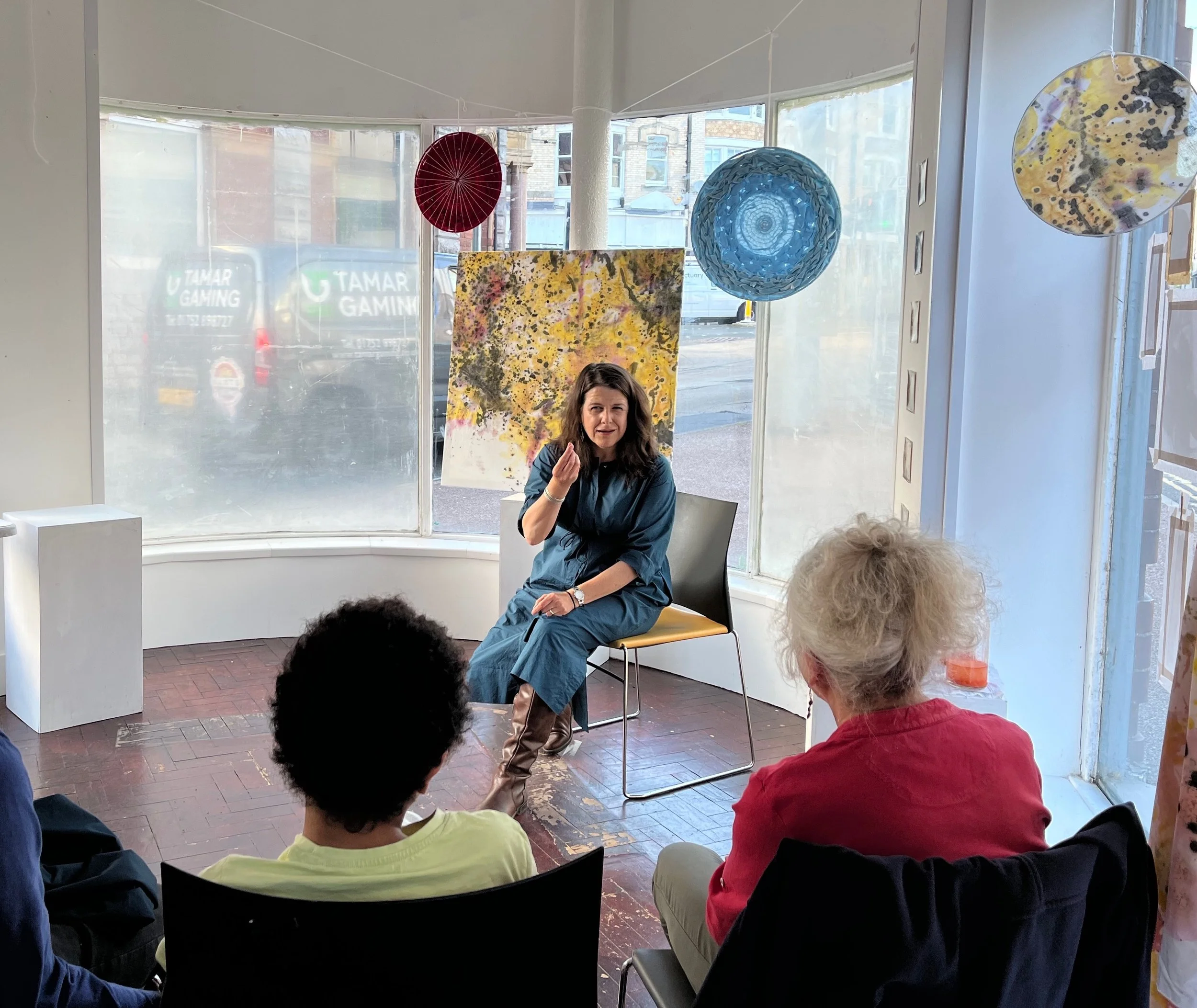 Lisa Schneidau is sitting on a chair, speaking during an event . There are four audience members, two with dark hair and bright clothing, seated and facing her. The scene is inside a room with large windows showing a city street outside.