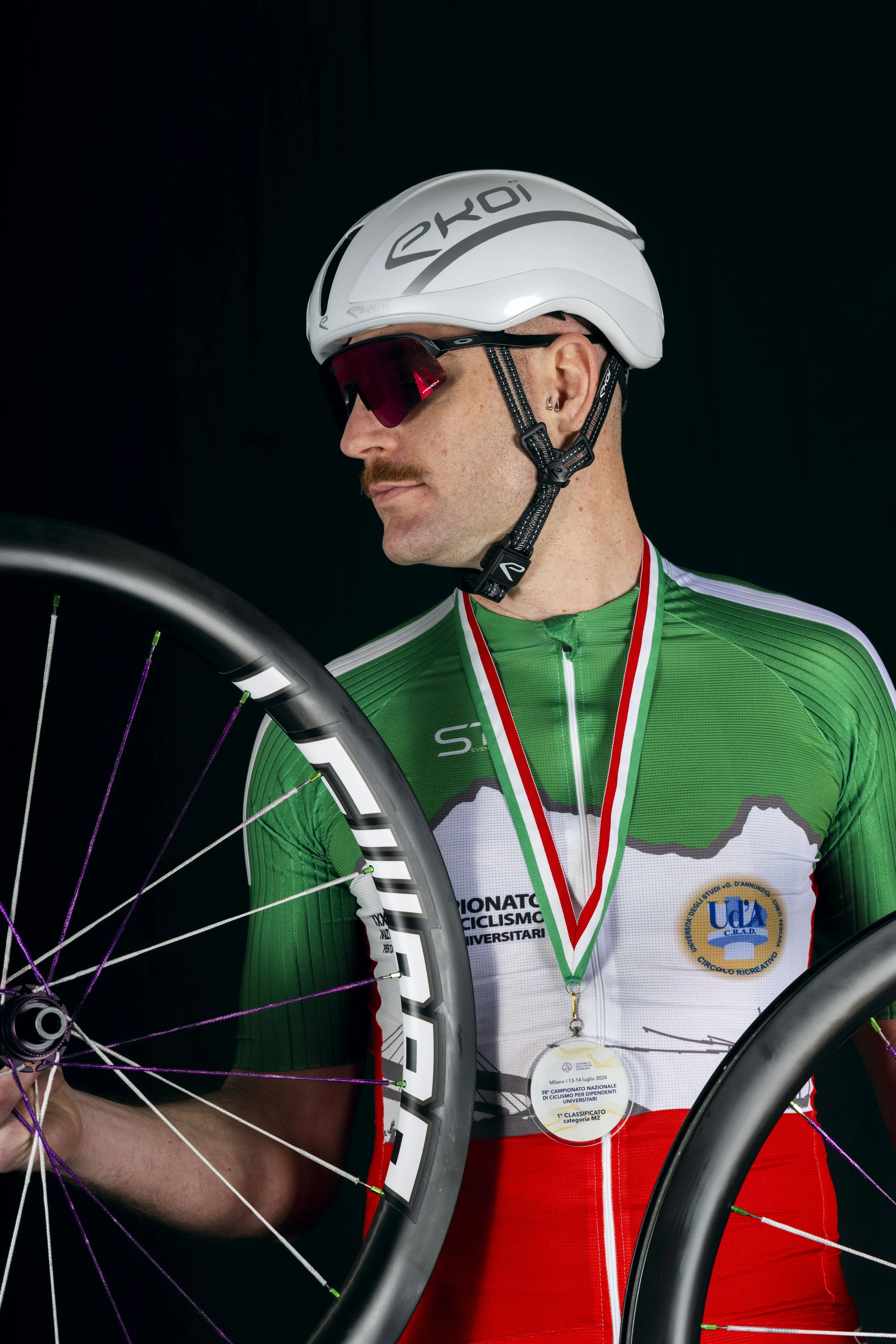 A cyclist in a green, white, and red jersey wearing a white helmet and sunglasses, holding a wheel, with a medal around his neck.