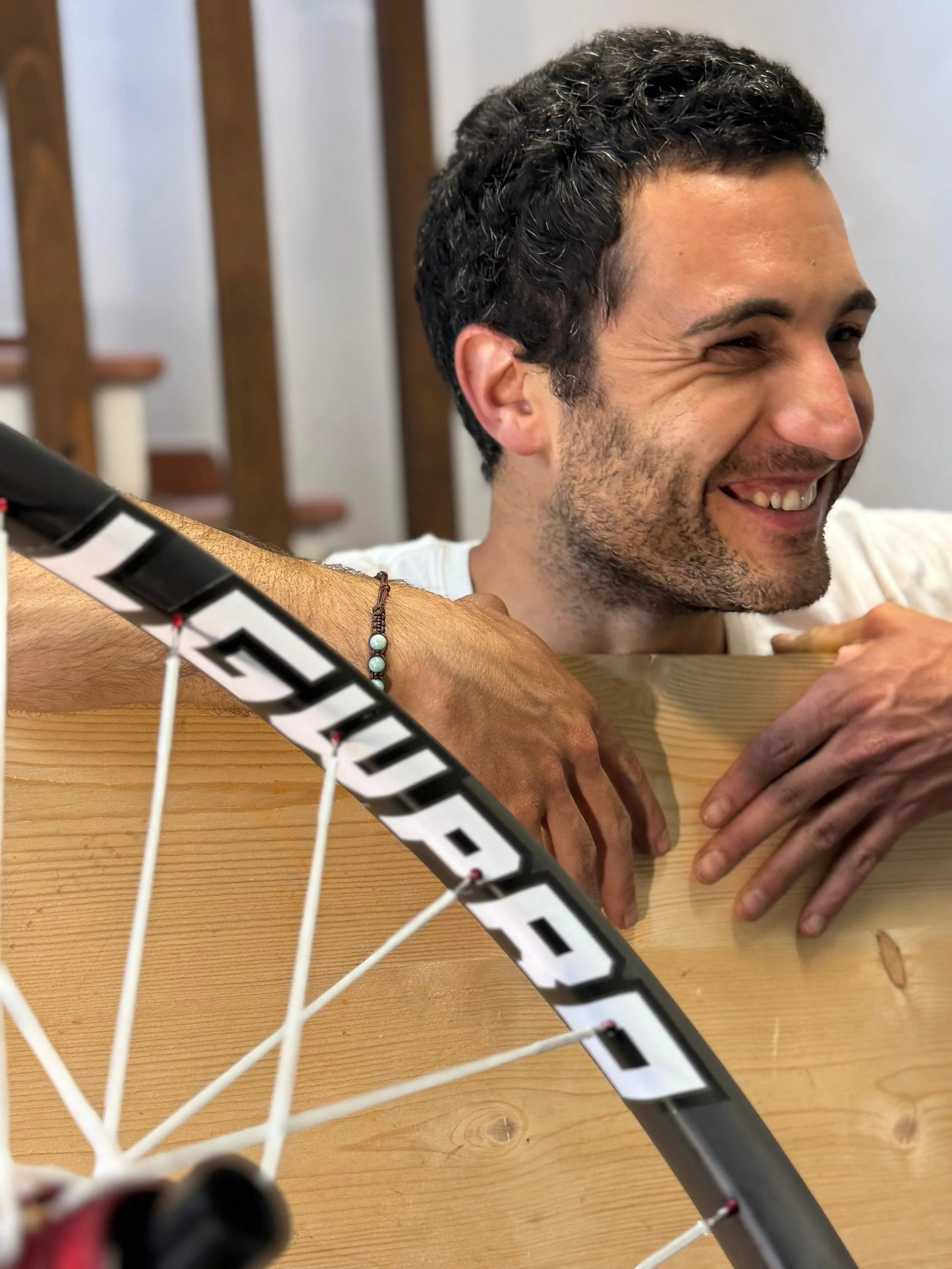 Uomo sorridente con mani appoggiate su un tavolo di legno, accanto a una bicicletta con ruota visibile in primo piano.