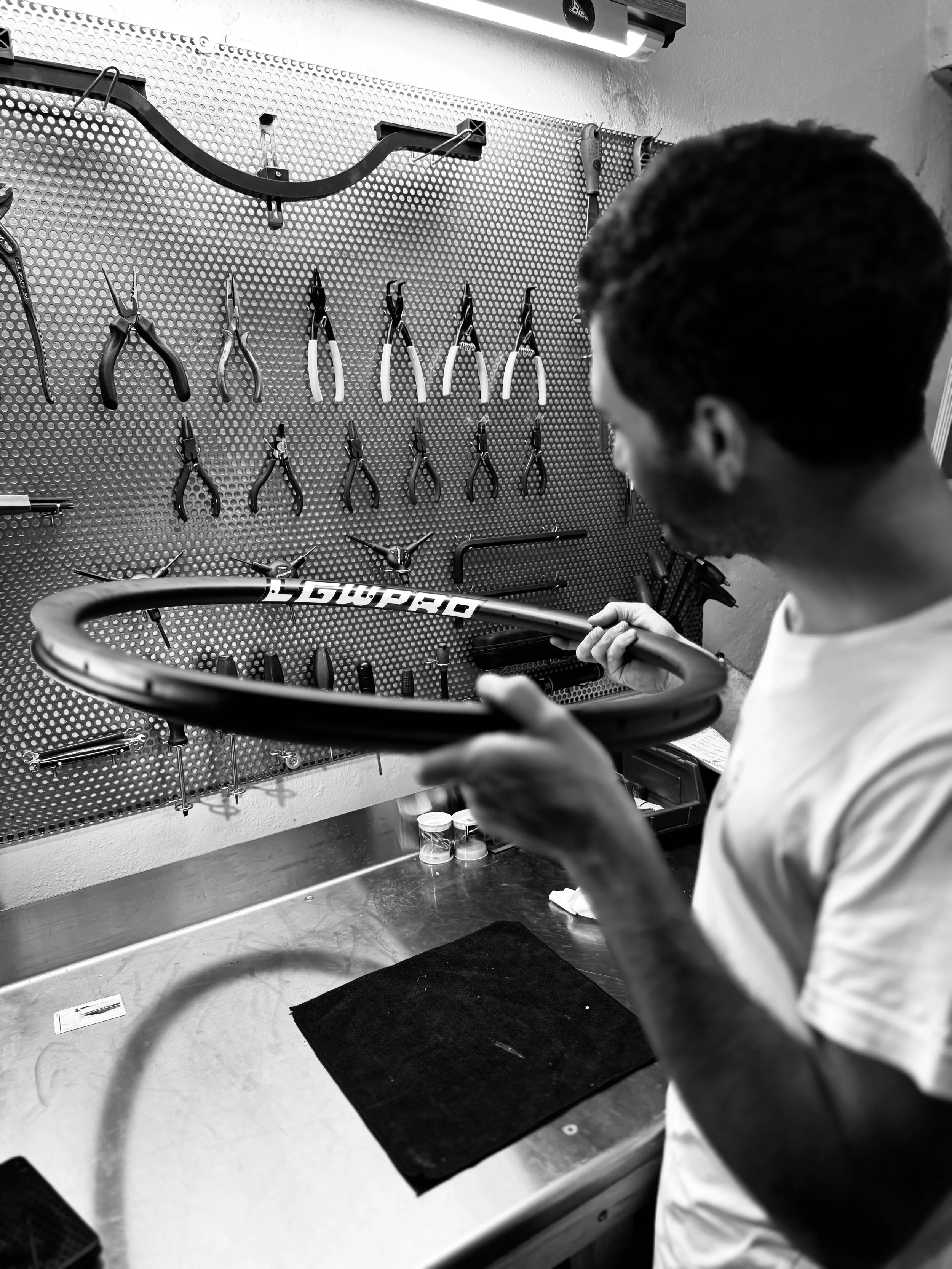 Un uomo sta ispezionando o riparando un cerchio di bicicletta in un'officina, con attrezzi appesi su una parete di pannello forato.