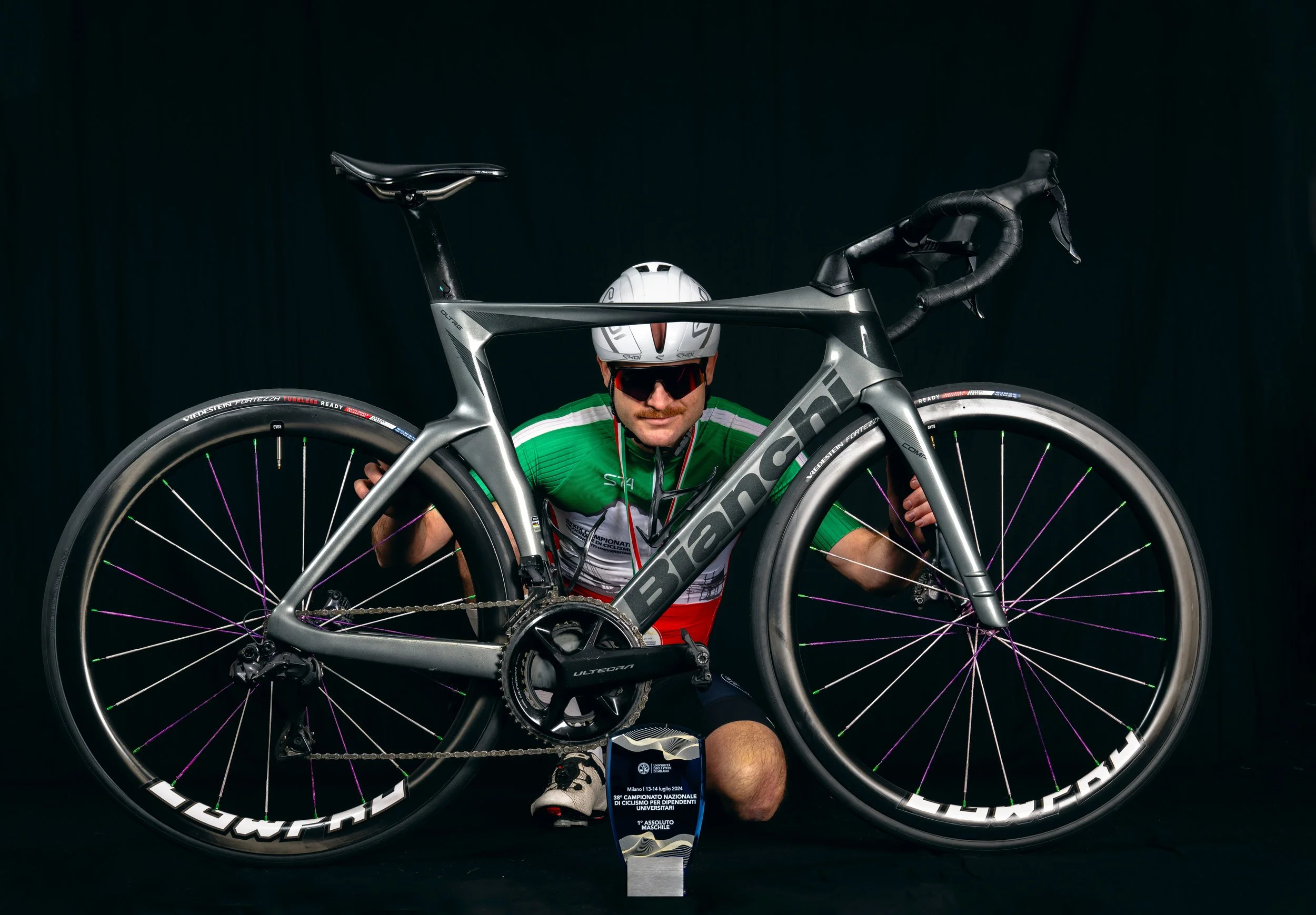 Uomo in abbigliamento da ciclismo con casco e occhiali da sole, accanto a una bicicletta da corsa, posato su un pavimento nero contro sfondo nero, con un trofeo davanti.