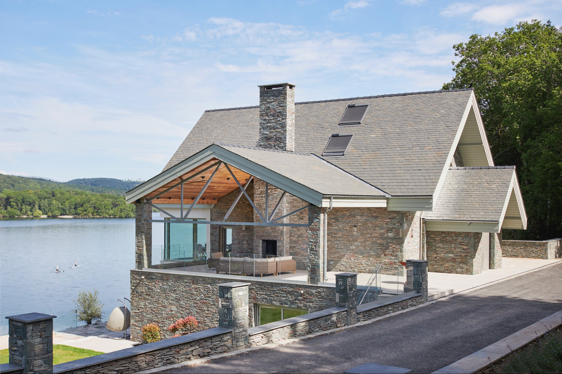Modern lakeside house with stone exterior, large glass windows, a screened porch, and a scenic view of water and green landscape.