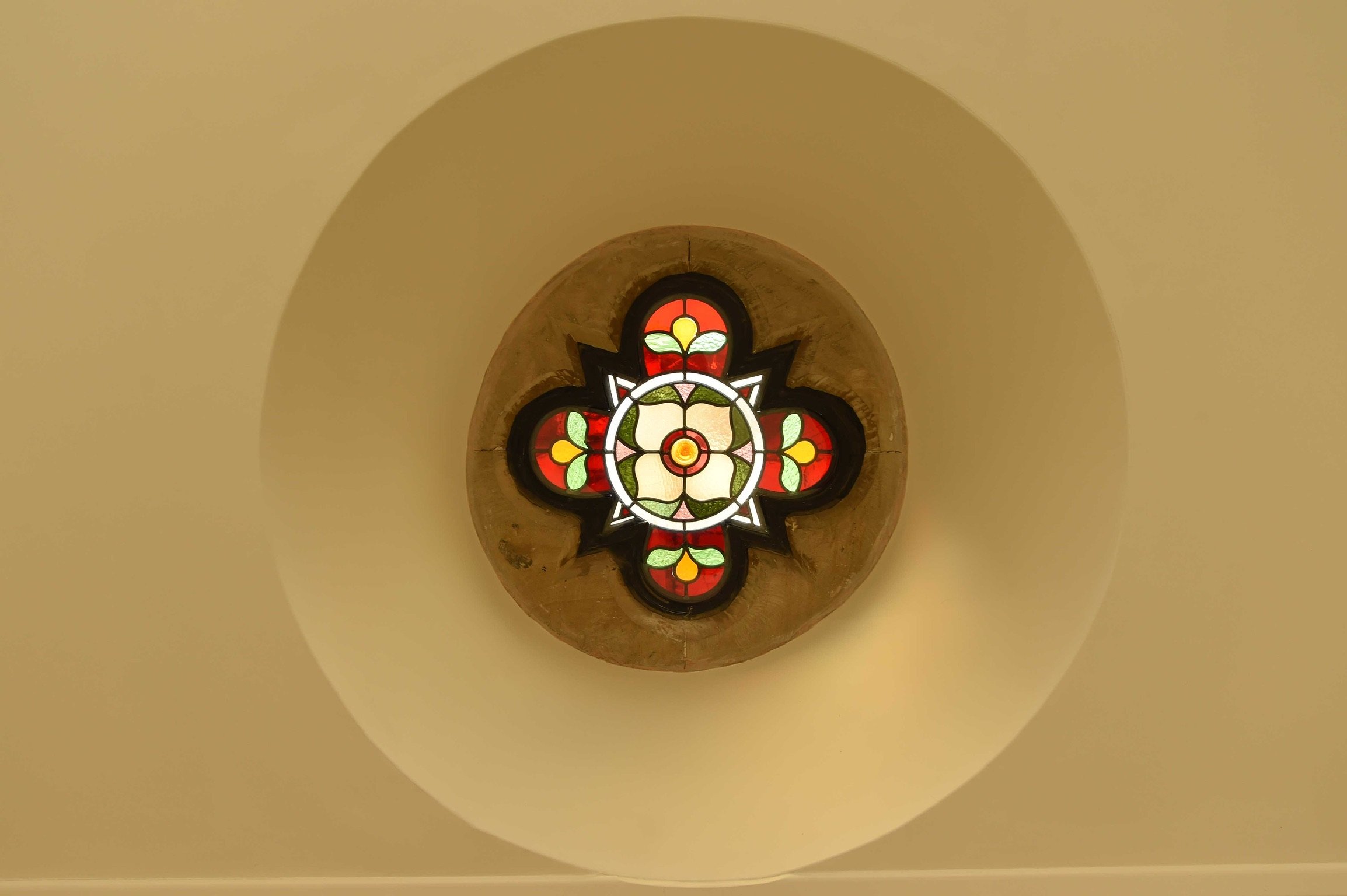Colorful stained glass window with red, yellow, green, and white patterns in a circular design, viewed from below against a beige background.