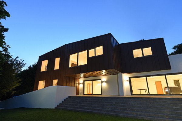 Modern two-story house with black and white exterior, illuminated windows and exterior lighting, with stairs leading to the entrance, surrounded by greenery at dusk.