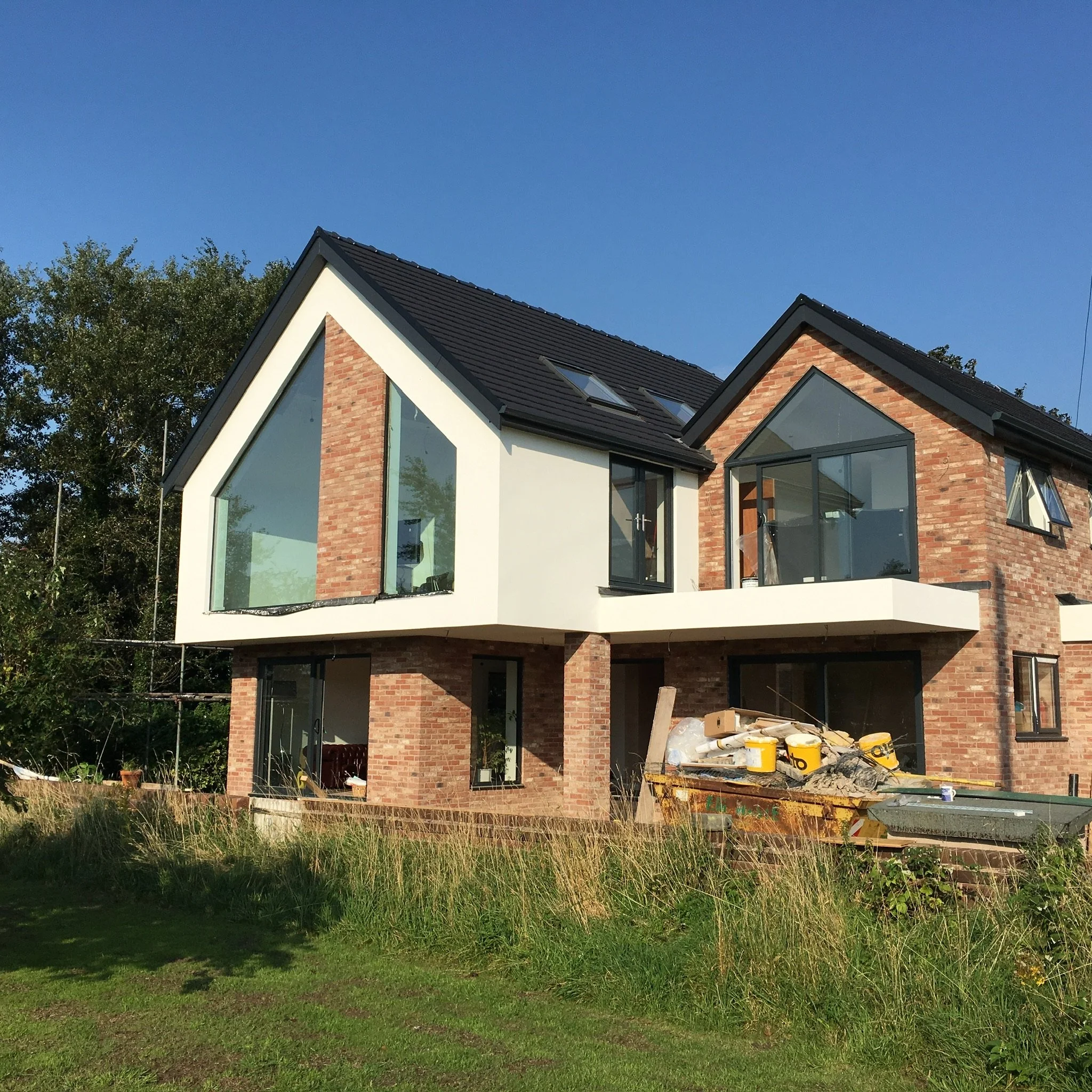 Modern two-story house under construction with brick and white exterior, large glass windows, and a black shingle roof, amid a grassy yard with construction materials in the foreground.