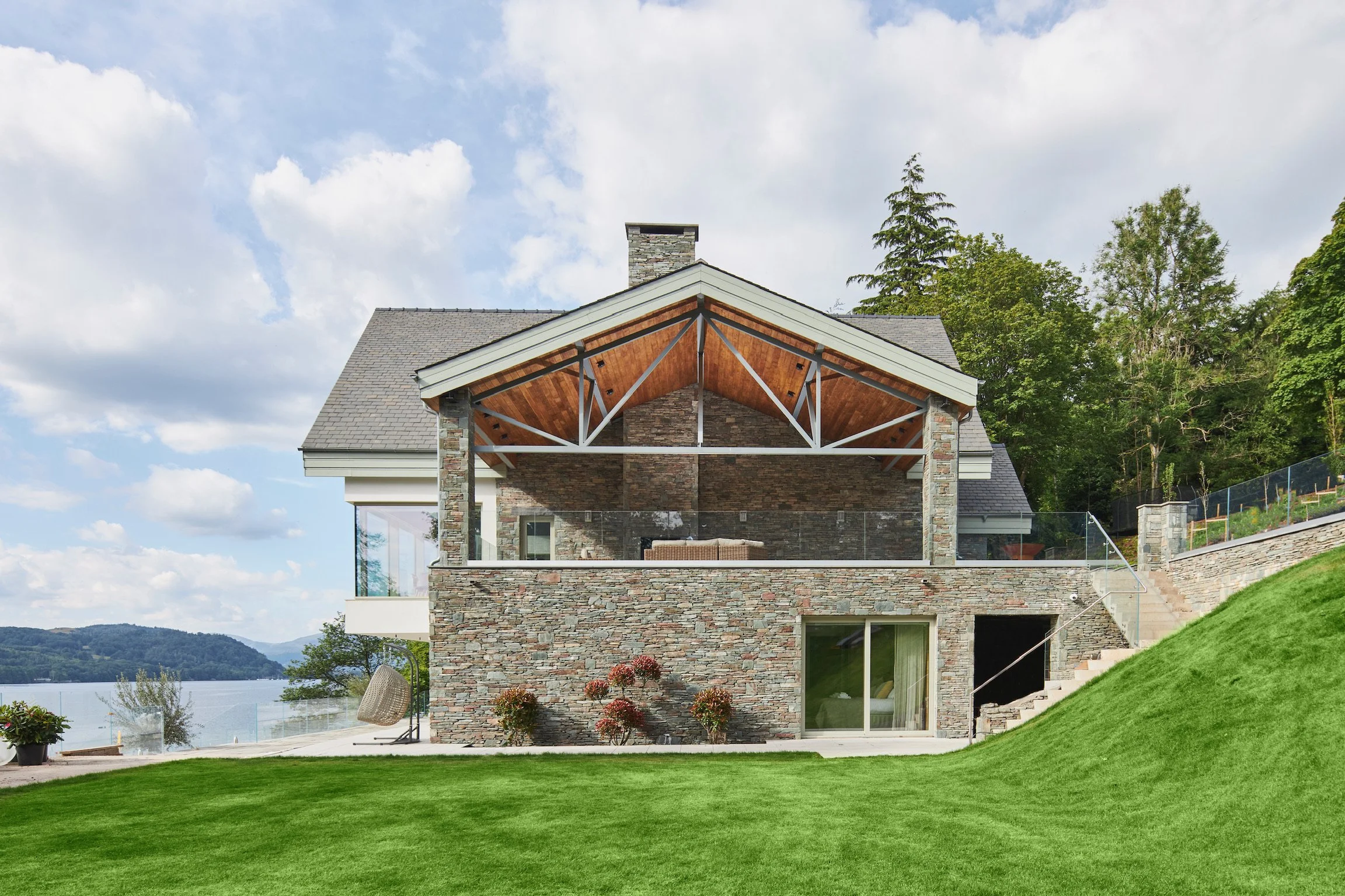 Modern house with stone and white exterior, large glass windows, and a wooden roof over a balcony, with a well-manicured lawn and trees in the background.