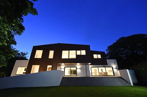Modern house with large windows and a dark and white exterior, illuminated at dusk, with stairs leading up to the entrance and trees surrounding it.