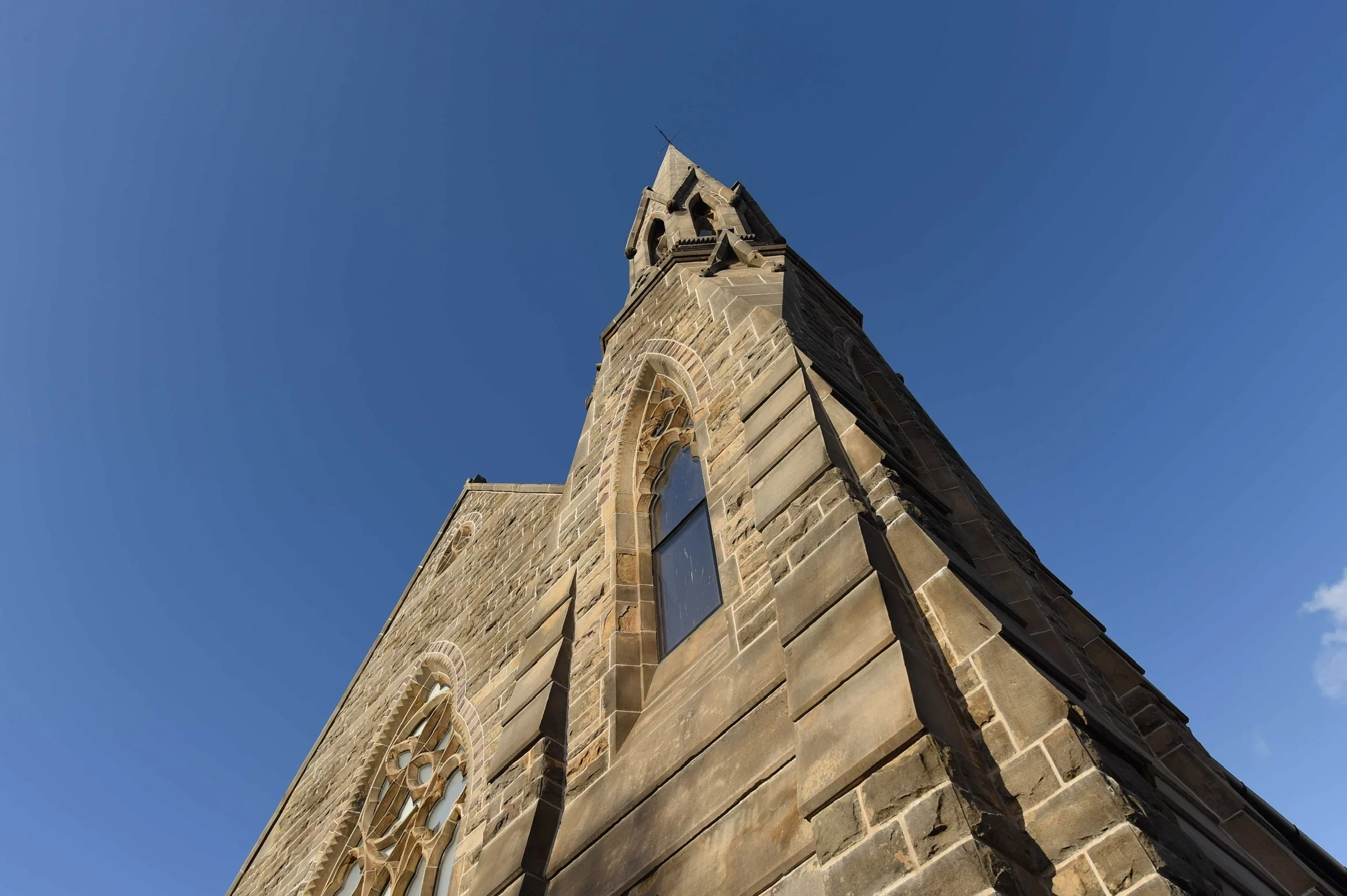Low-angle view of a stone church tower with arch windows and a spire against a clear blue sky.