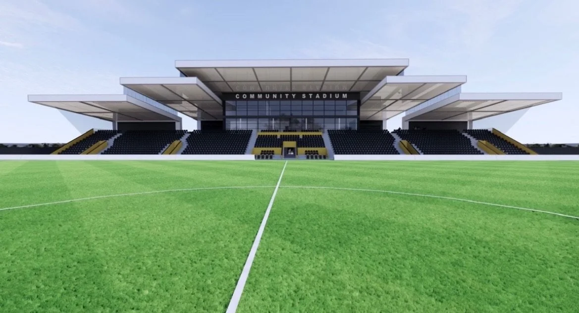 Modern community stadium with a green soccer field in the foreground, black seating, and large overhanging roof structures.