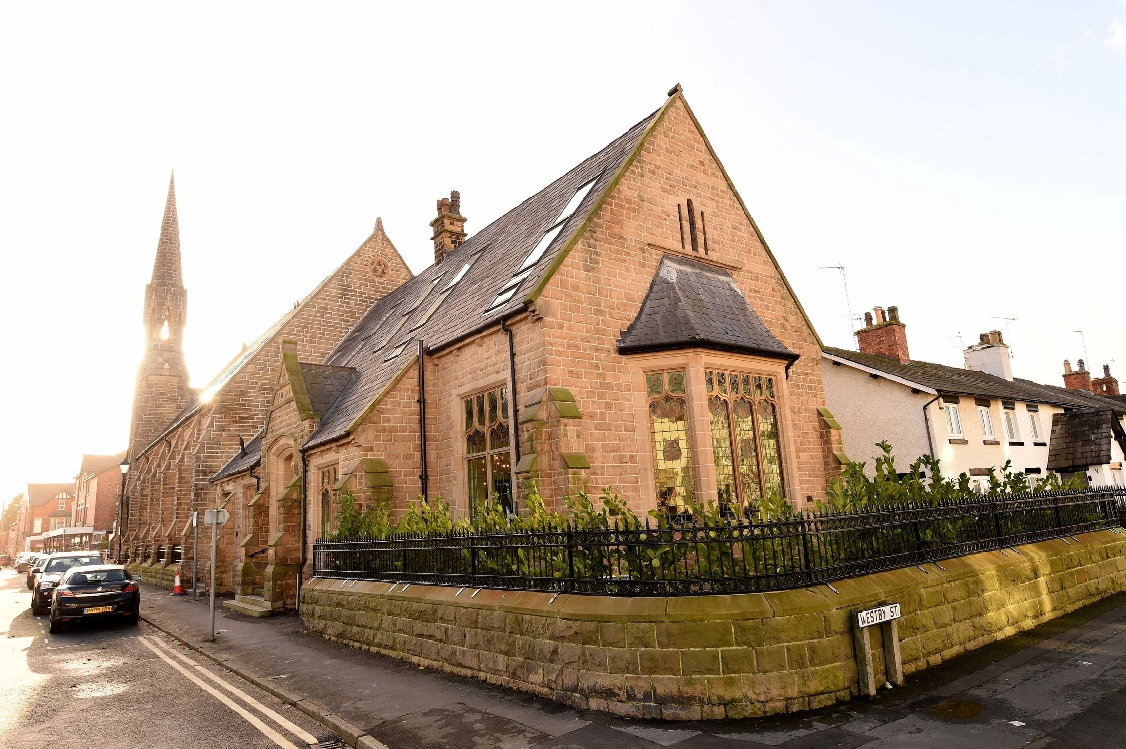 A small historic church made of red bricks with large stained glass windows, located on Westby Street, with neighboring houses and a church steeple visible in the background, bathed in warm sunlight.