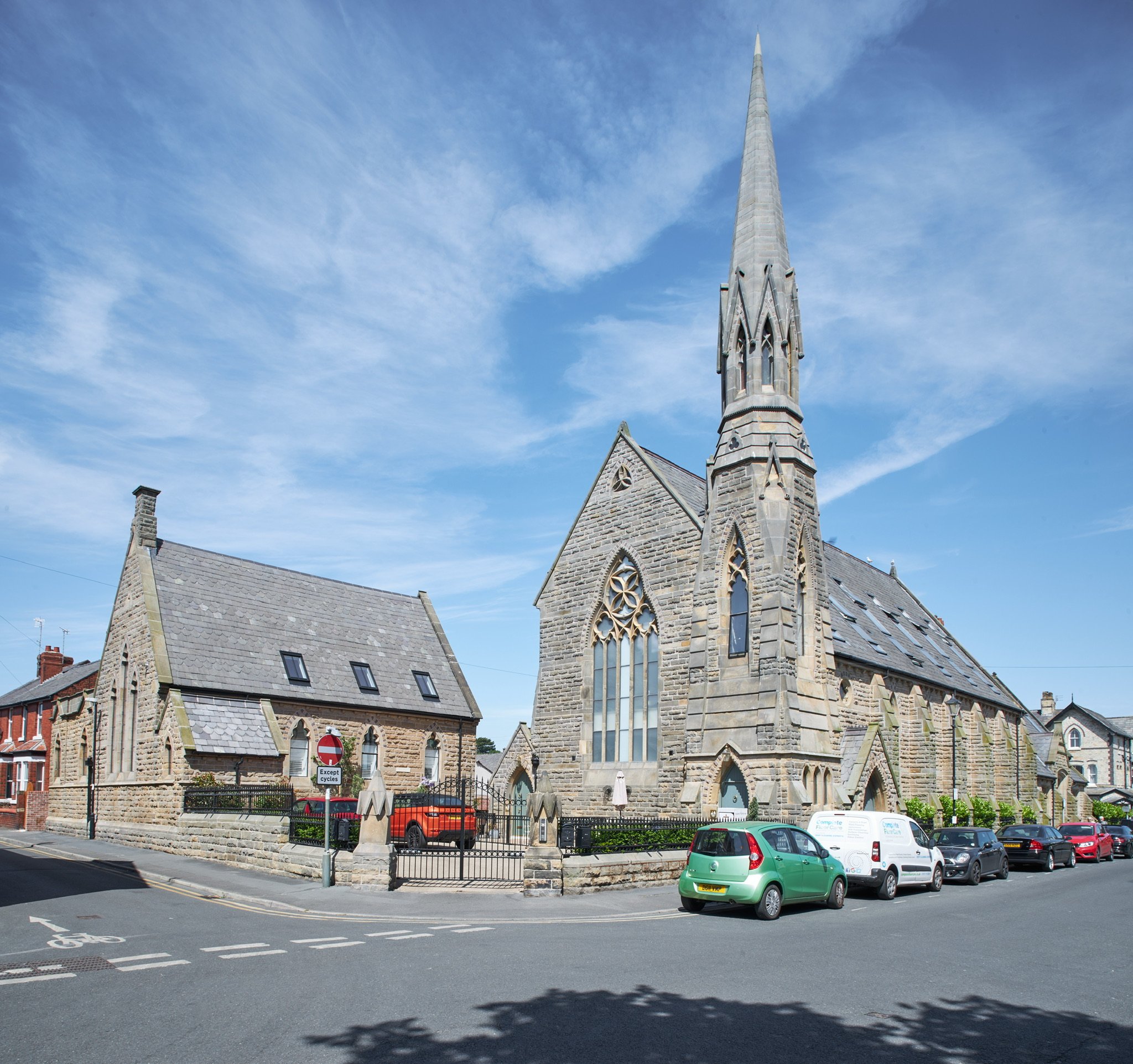 A historic stone church with a tall steeple, large stained glass windows, and adjacent buildings, surrounded by parked cars under a partly cloudy blue sky.