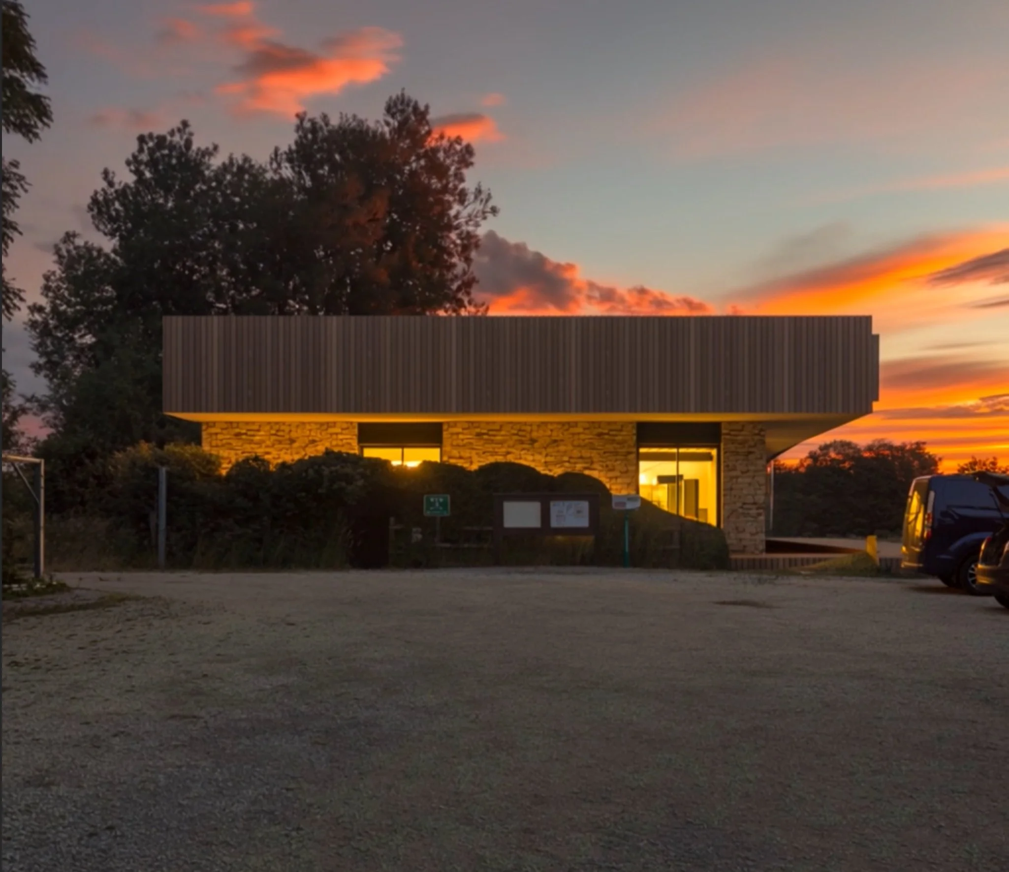 A modern building with a curved roof, stone and metal exterior panels, illuminated windows, and a dirt parking lot in front at sunset with colorful clouds and trees in the background.