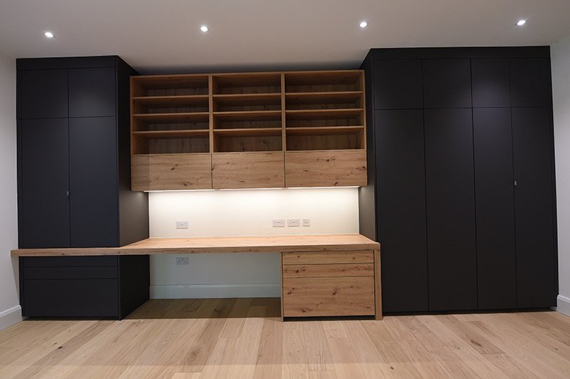 Modern built-in office or storage unit with black cabinets on each side, wooden shelves and desk, and a white wall with electrical outlets in the background.