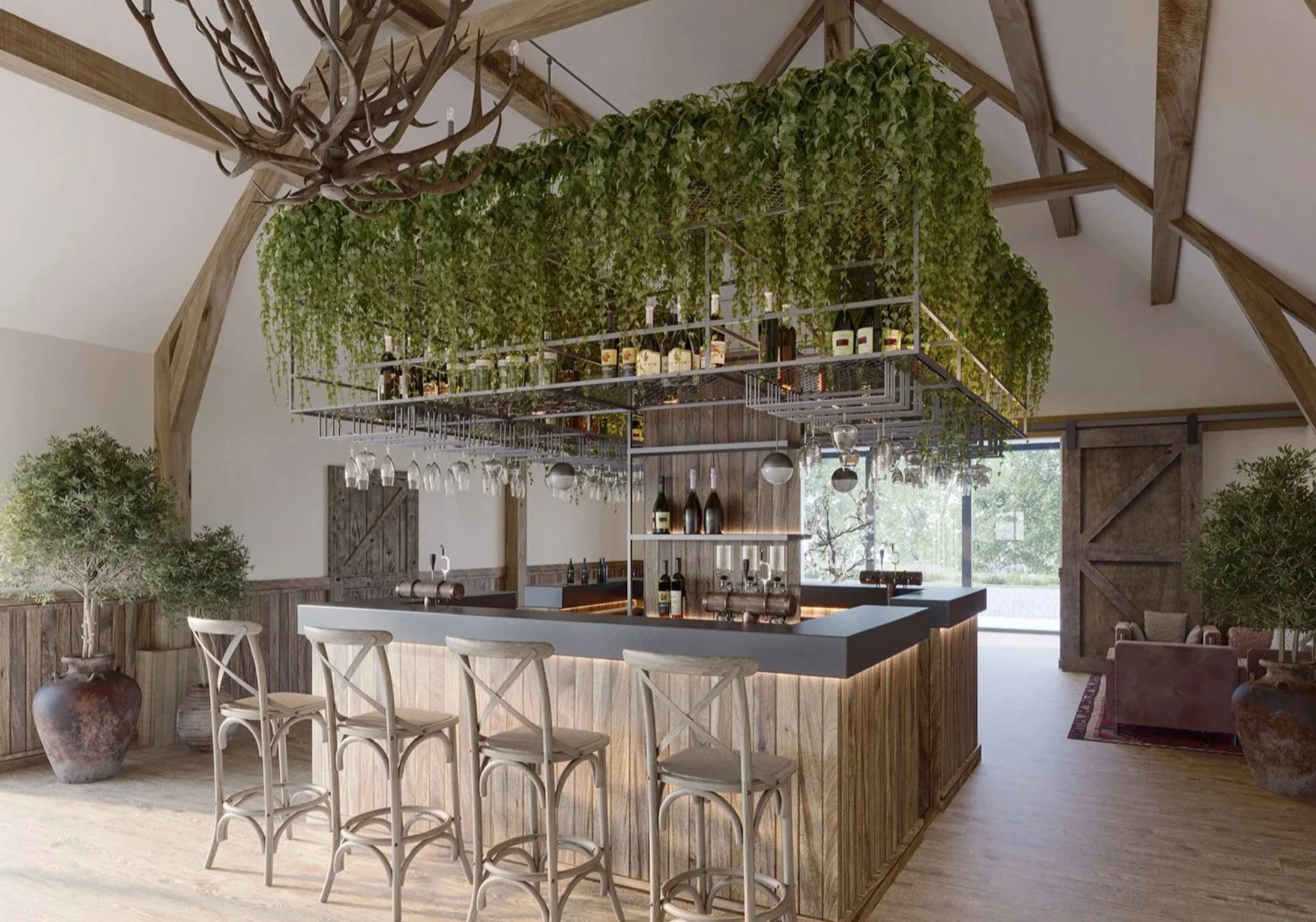 A rustic indoor bar with a wooden counter and four chairs. Above the bar, a metal rack holds bottles, glasses, and greenery. A large antler chandelier hangs from the ceiling. Potted plants are on the sides, and a sliding barn door is in the backgroun