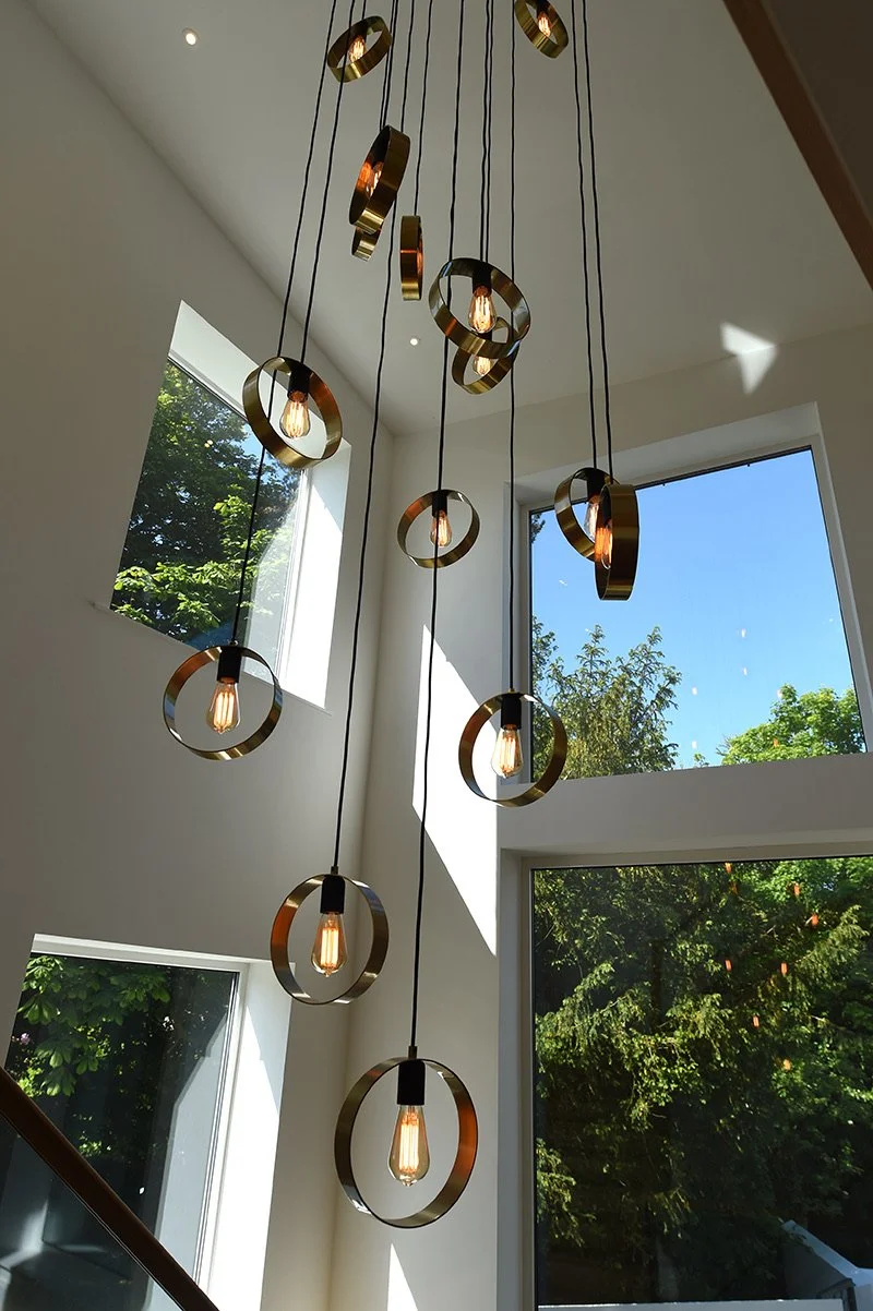 Modern chandelier with multiple hanging lights inside a bright room with large windows showing green trees outside.