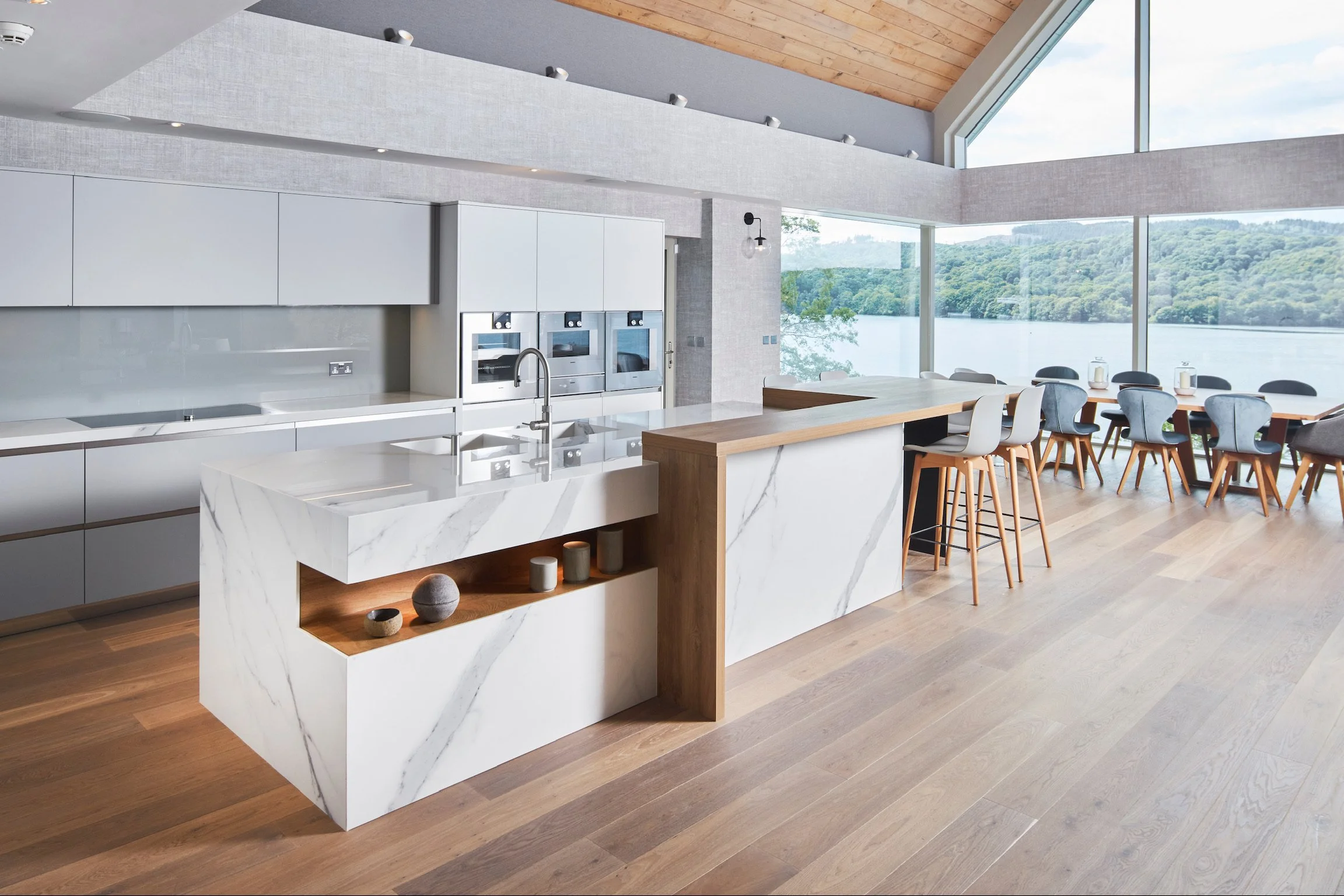 Modern kitchen with marble island, white cabinets, built-in ovens, wooden bar stools, and large windows overlooking a lake and trees.
