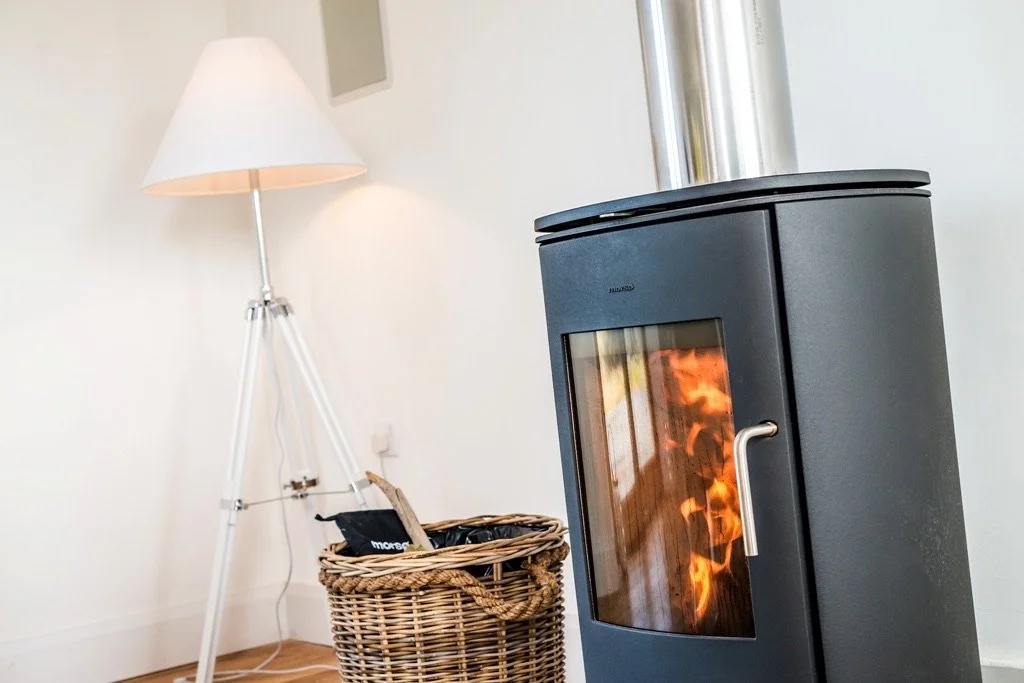 A black pellet stove with a glass door showing visible flames inside, positioned in a room with a wicker basket beside it, a white floor lamp with a tripod base behind, and a white wall with a power outlet.