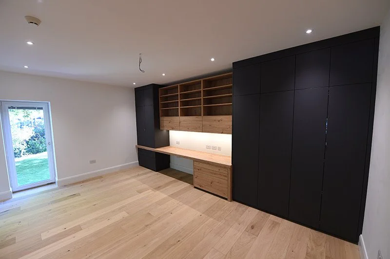 Empty room with light wooden floor, white walls, built-in wooden desk and shelving unit, and black storage cabinets. A glass door leading outside, with greenery visible.