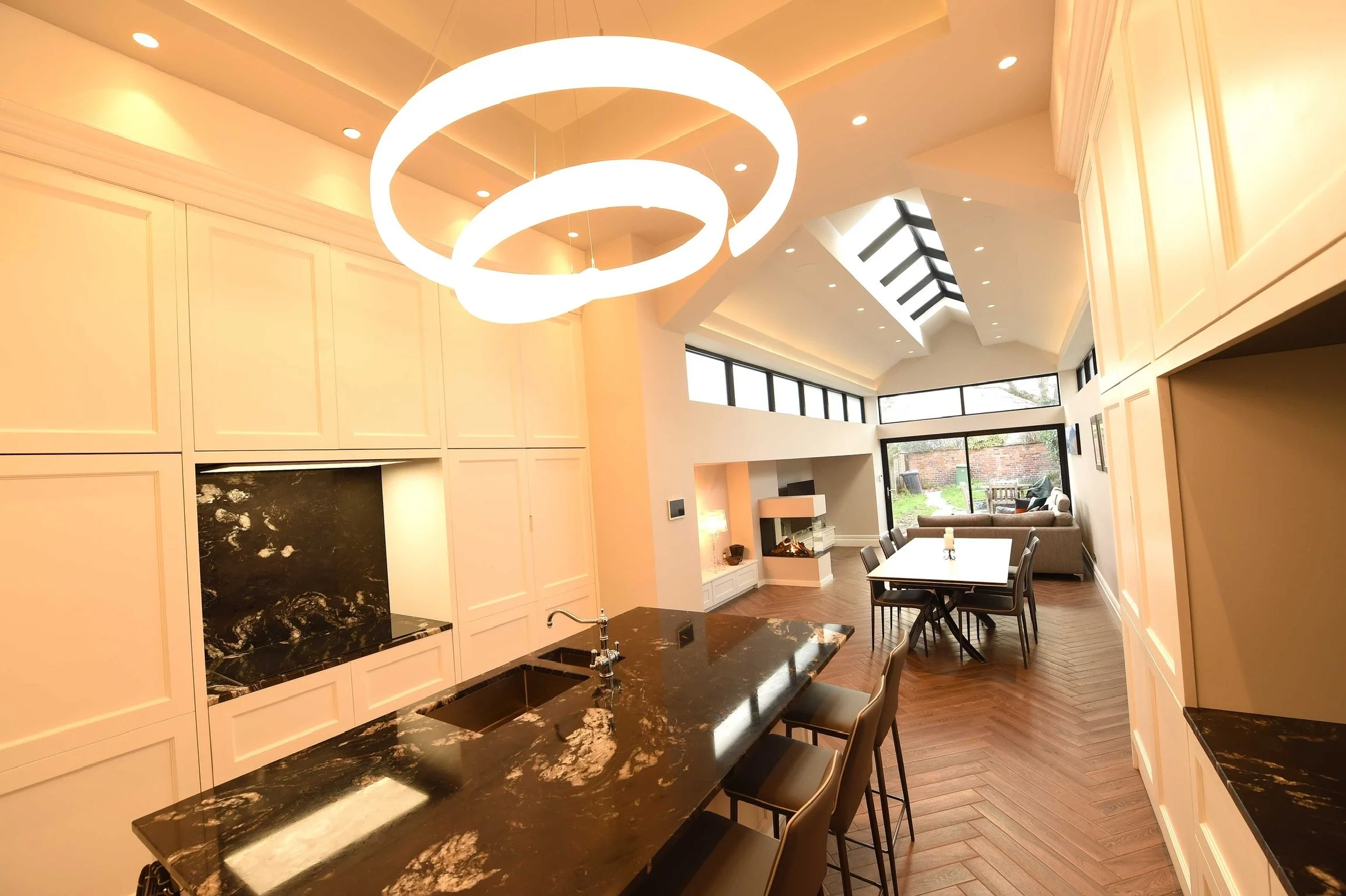 Modern open-concept living space with kitchen and dining area, featuring black marble countertops, a living room with a fireplace, and large windows bringing in natural light.