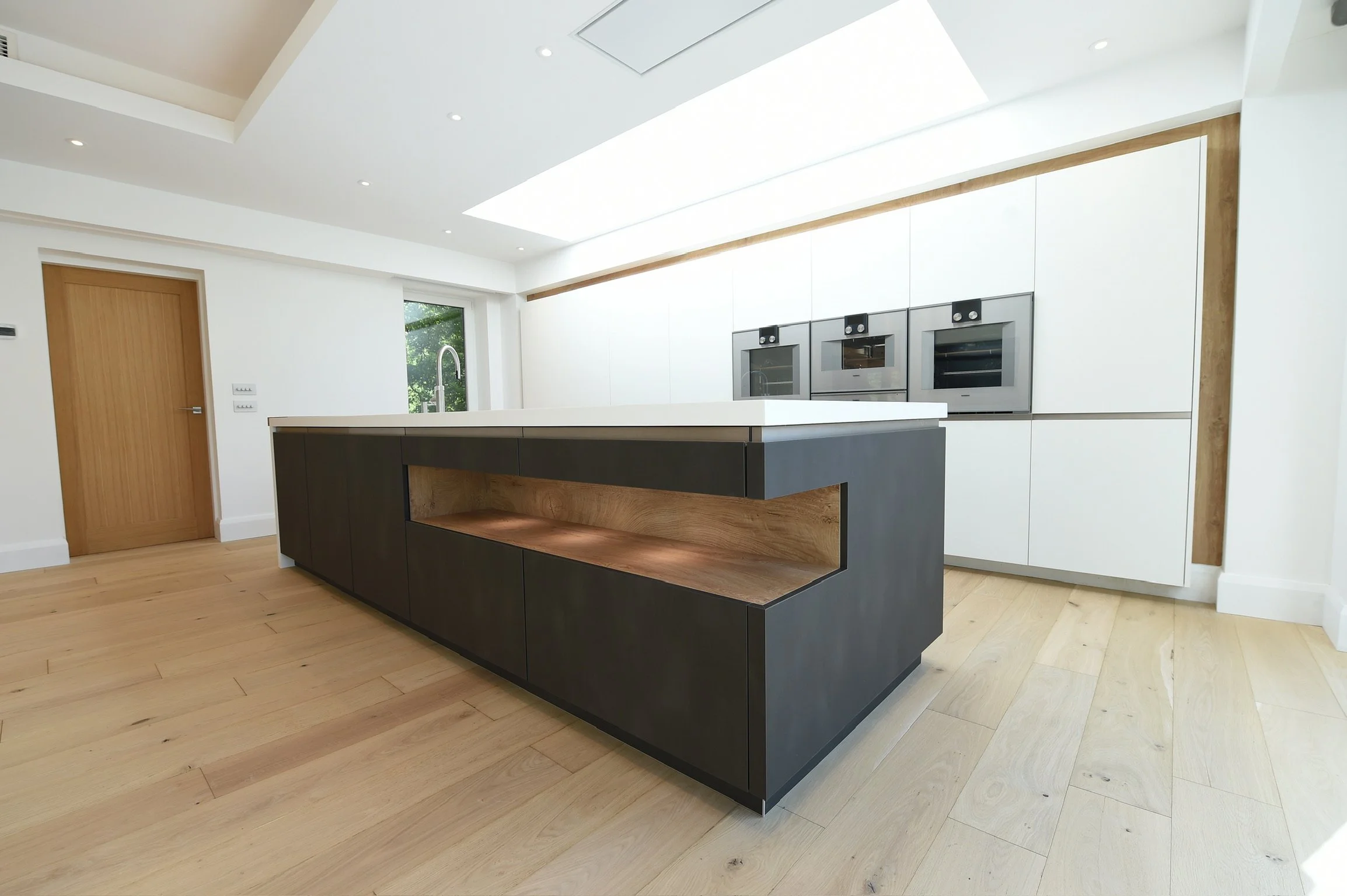 Modern kitchen with minimalist design featuring a black and wooden kitchen island, white cabinetry, a built-in oven, microwave, and a large window with tree view.