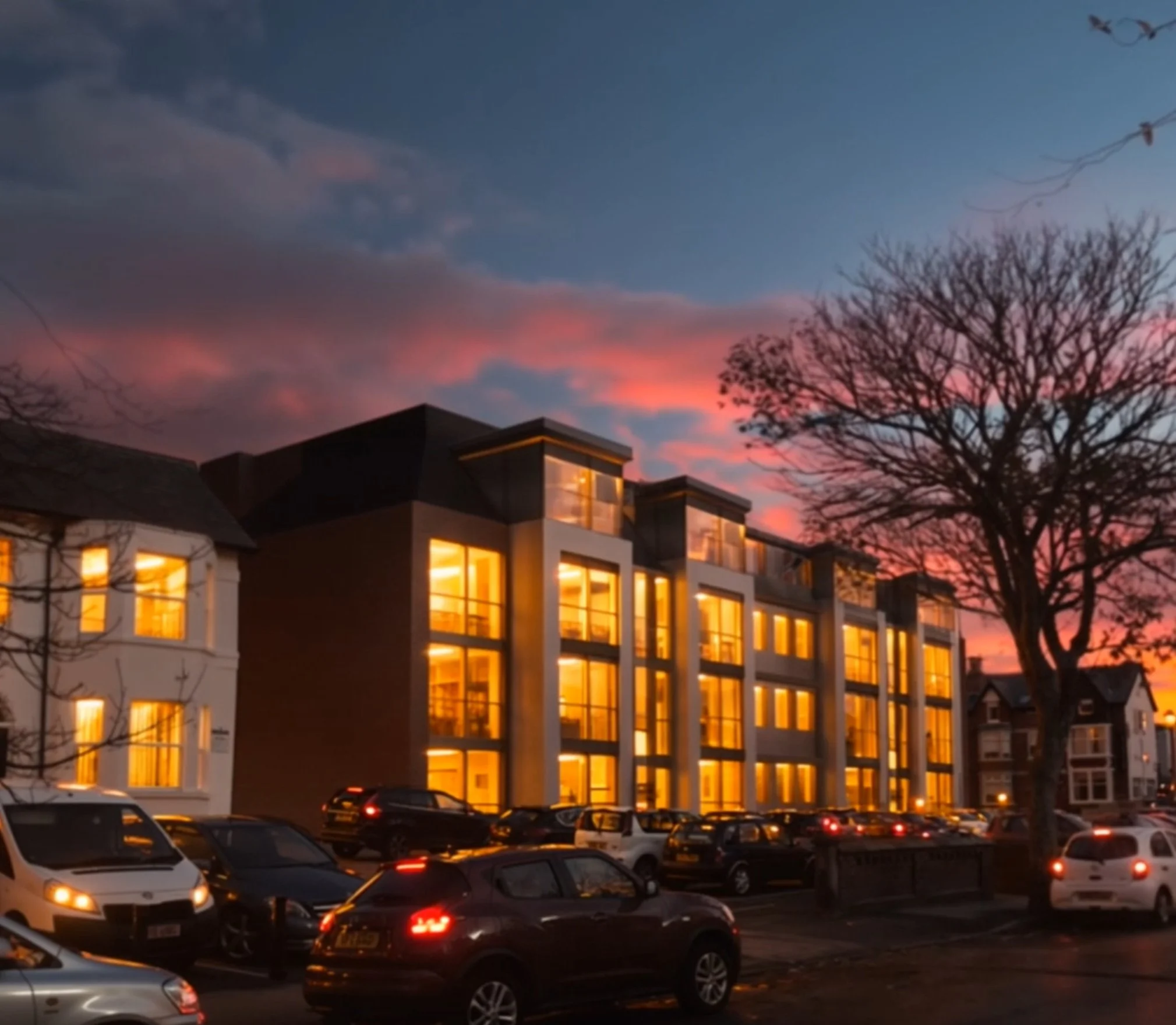 Several parked cars in front of a modern building with large, illuminated windows at sunset, with a leafless tree and a colorful sky in the background.
