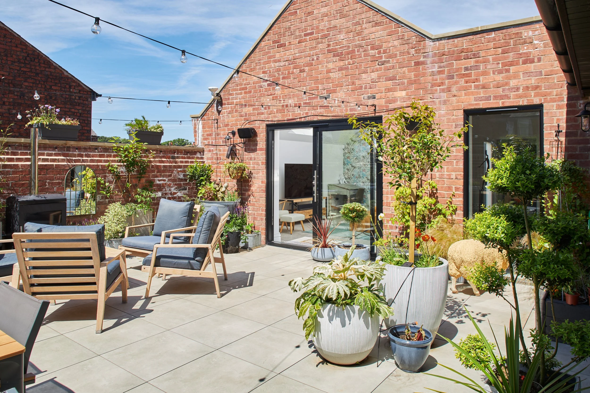 Sunny rooftop terrace with outdoor seating, potted plants, and string lights, adjacent to a brick building with sliding glass doors.