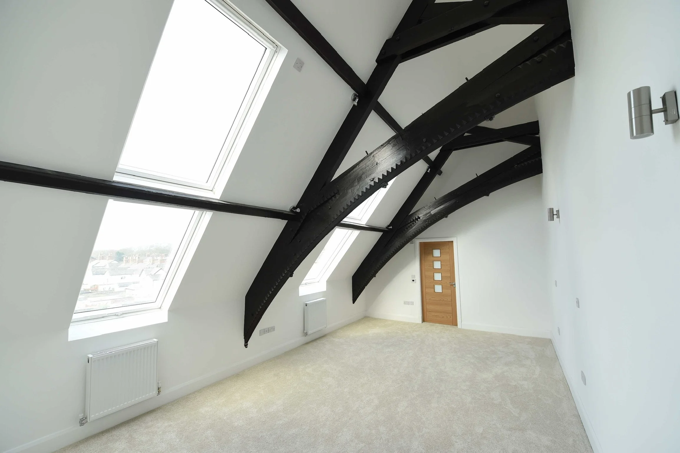 Empty, modern attic room with white walls, beige carpet, large skylight windows, black exposed wooden beams, and a wooden door.
