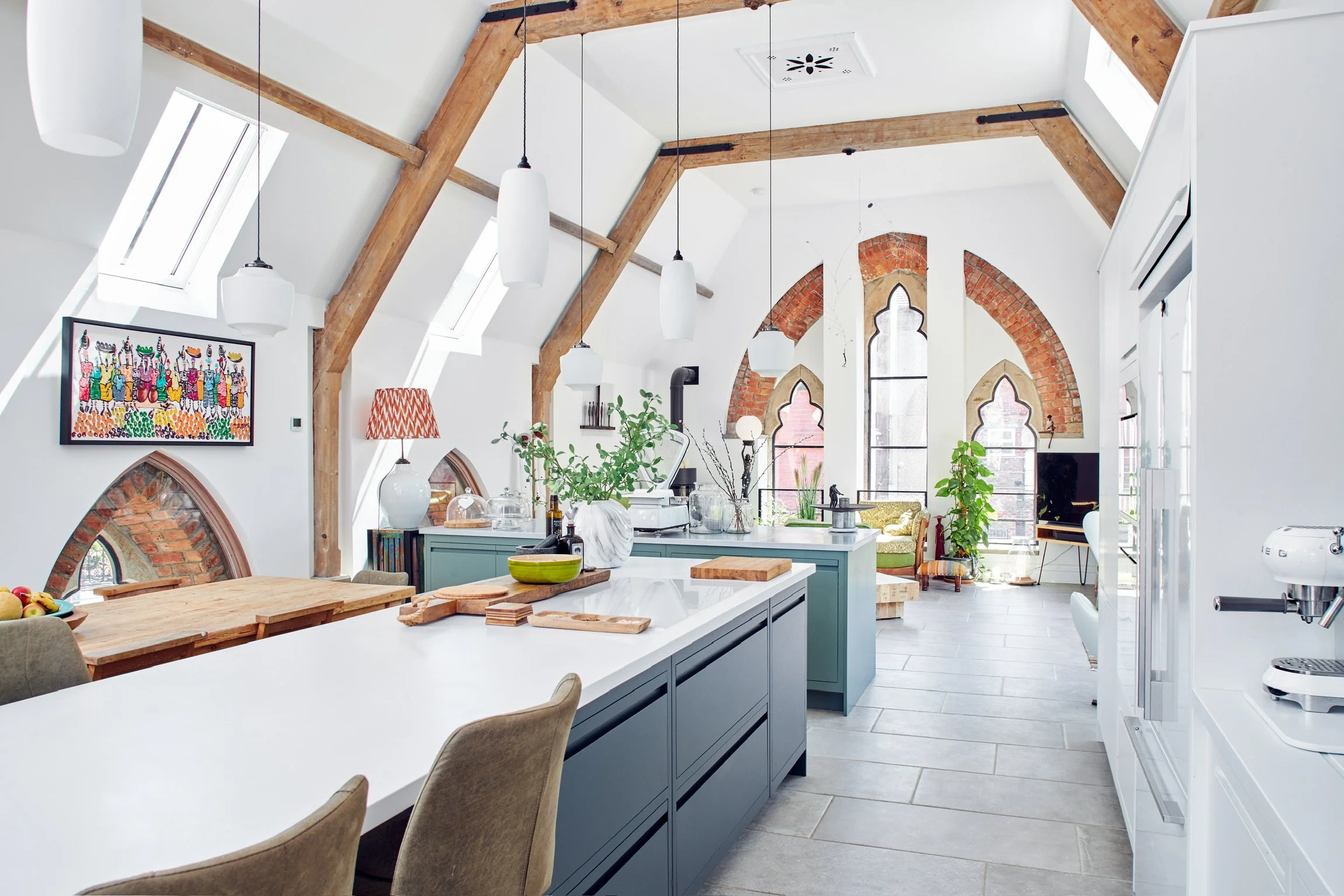 A bright and airy kitchen with white walls, large skylight windows, and exposed wooden beams. Features a kitchen island with a white countertop, blue cabinets, and various plants and decor items. In the background, there is a sitting area with a gree