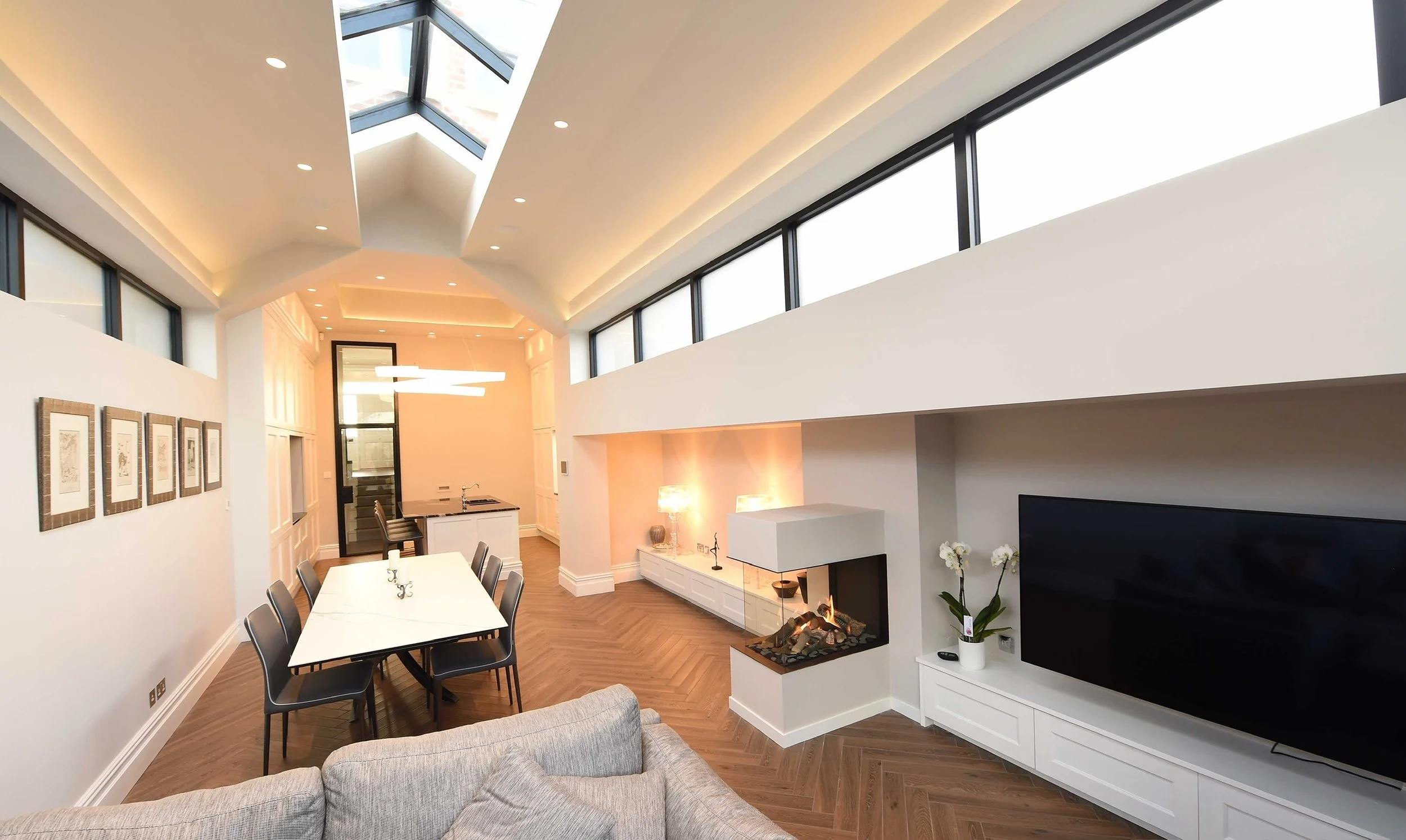 Modern living room with white walls, a fireplace, flat-screen TV, dining table with six chairs, artwork on wall, and large windows with a skylight.