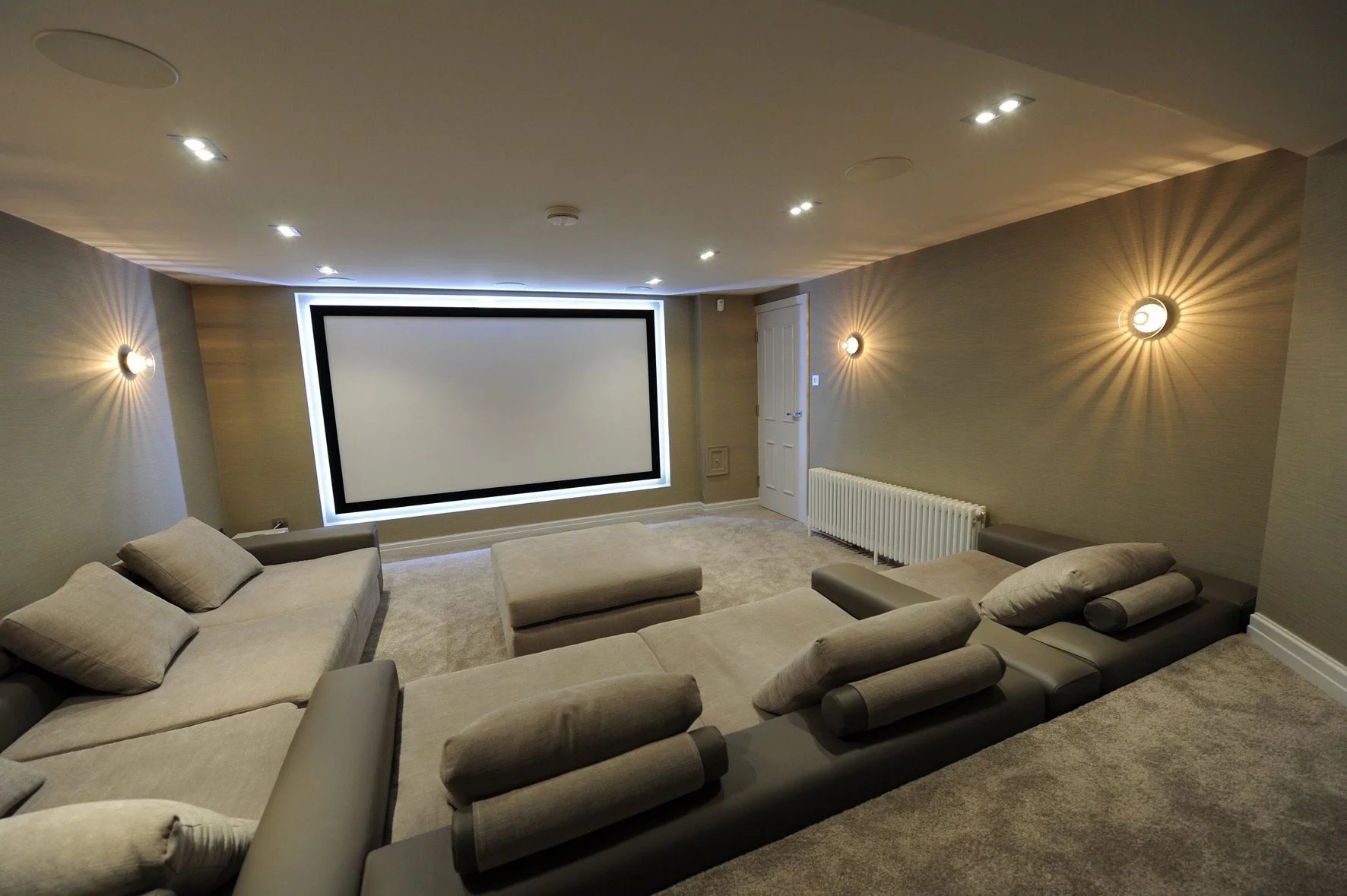 A modern home theater room with beige sofas, a large projection screen framed with backlit lighting, and warm wall-mounted lights creating a cozy ambiance.