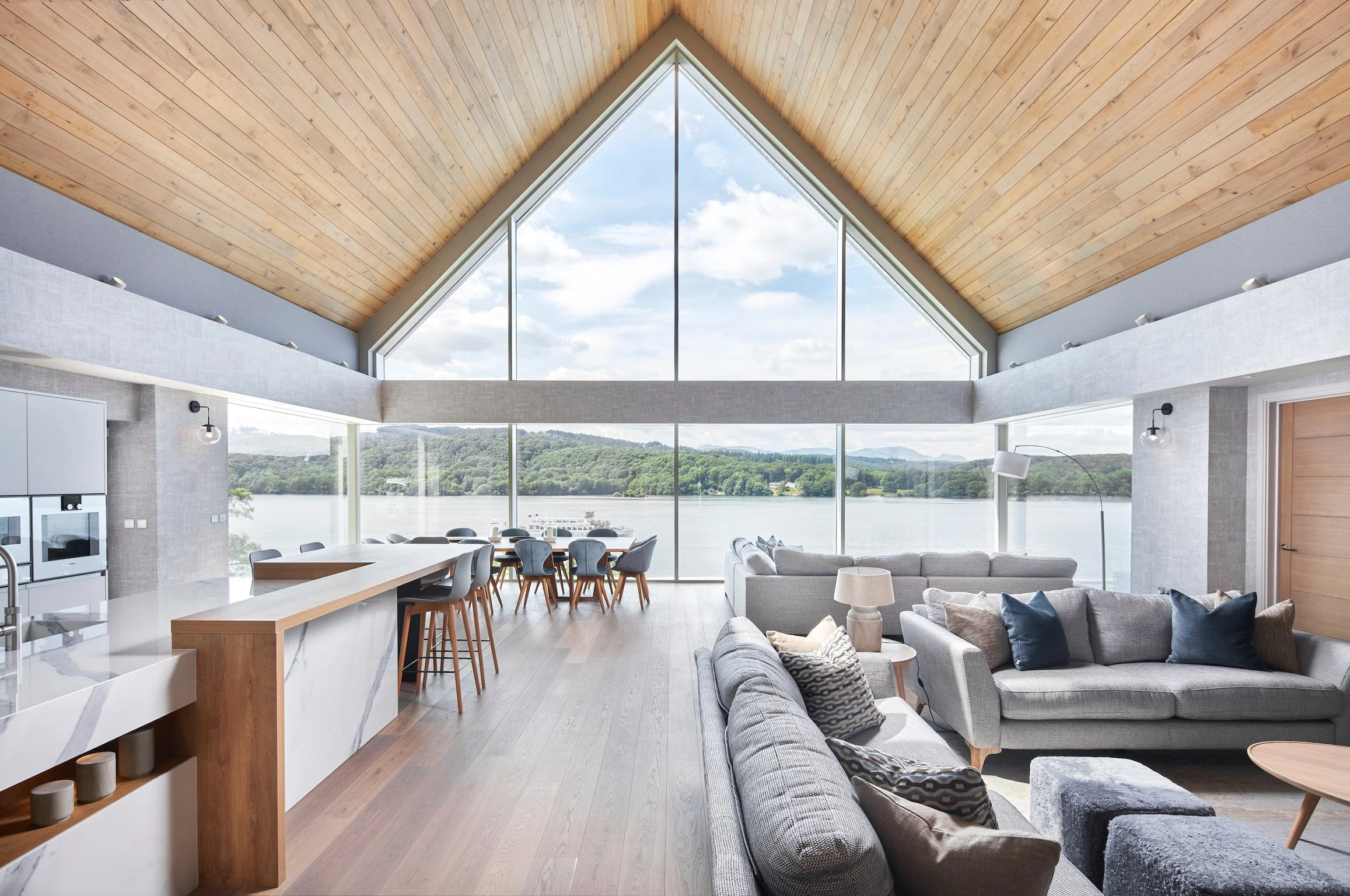 Living room and dining area with large floor-to-ceiling windows overlooking a lake and green hills, with a wooden vaulted ceiling and modern furniture.