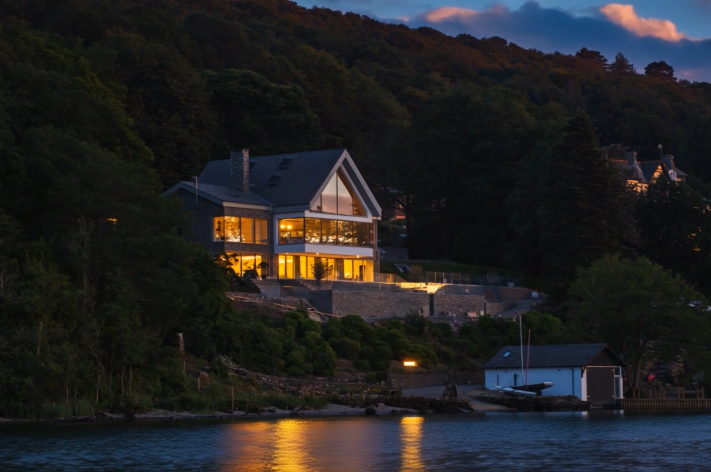 A modern house with large glass windows illuminated at dusk, situated on a hillside by a body of water, surrounded by trees and other houses in the distance.