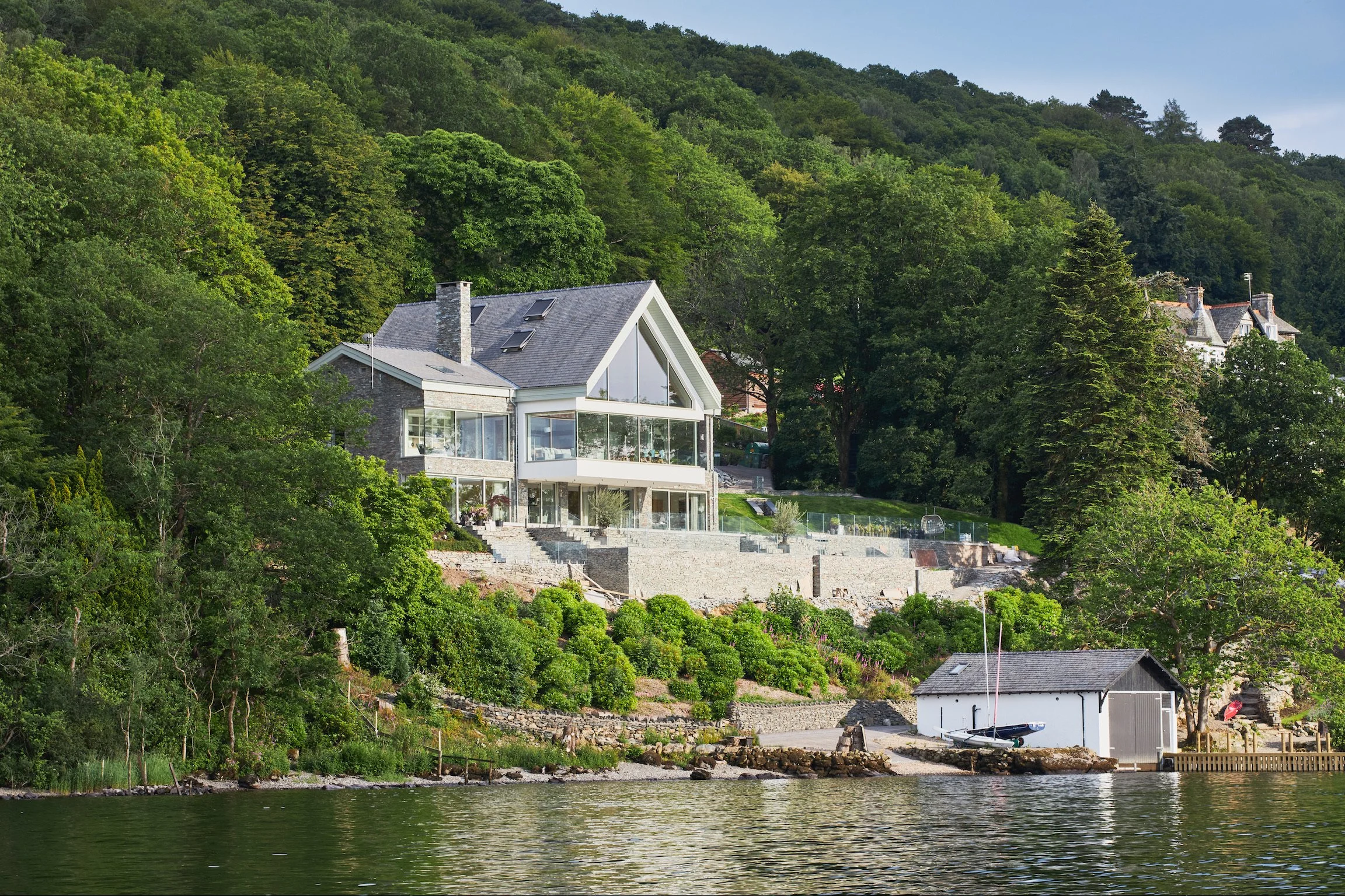 Modern house on a hillside by the water with trees and greenery around, and a small boat on the shore.