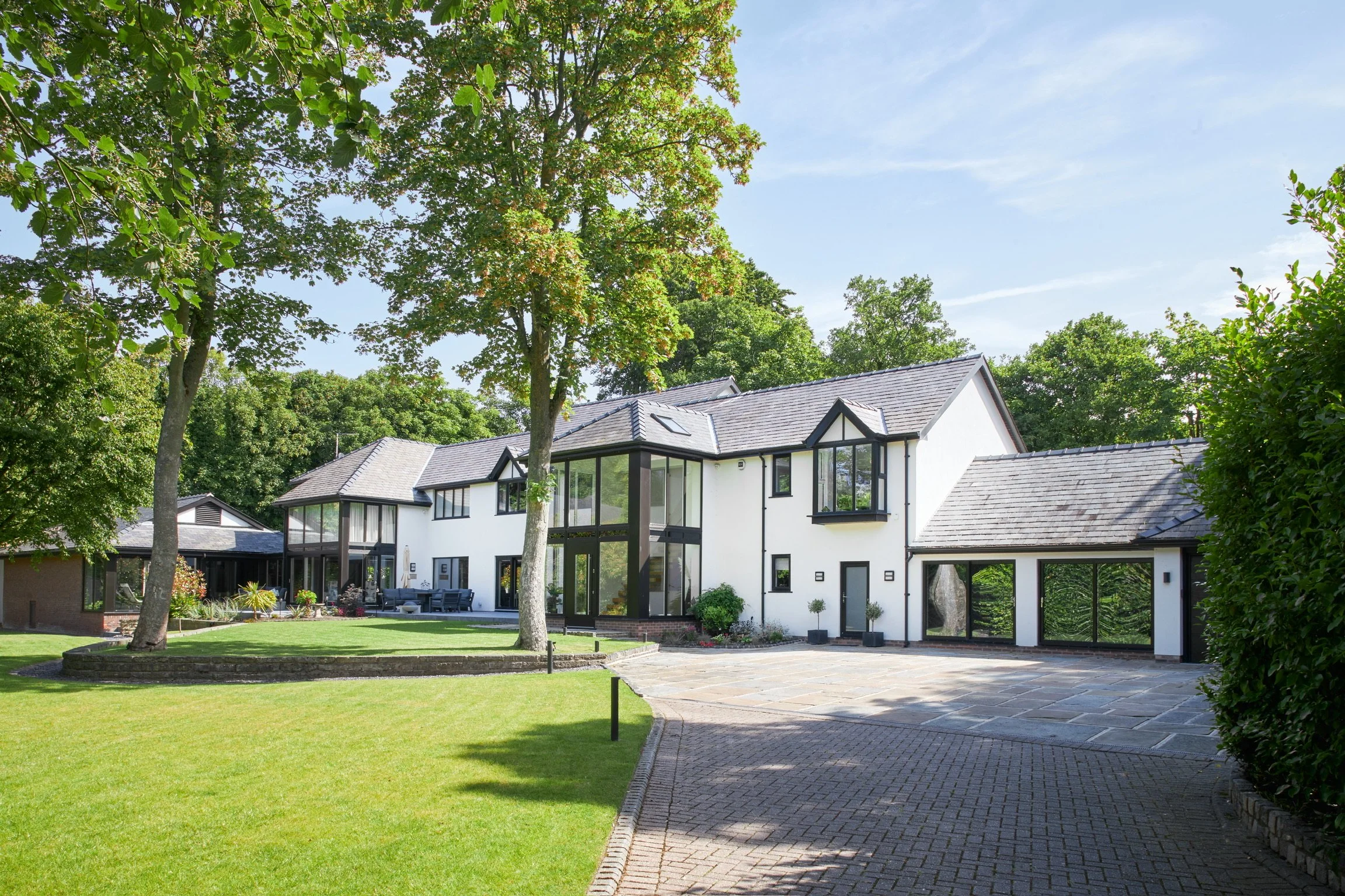 Modern white house with large glass windows, surrounded by green lawn and tall trees on a sunny day.